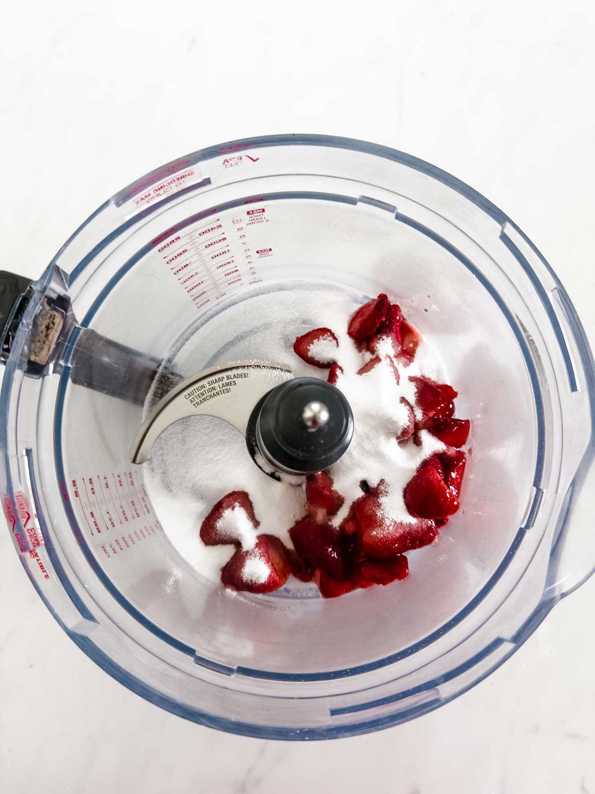 A food processor bowl containing granulated sugar and sliced strawberries, ready to be blended, on a white surface.