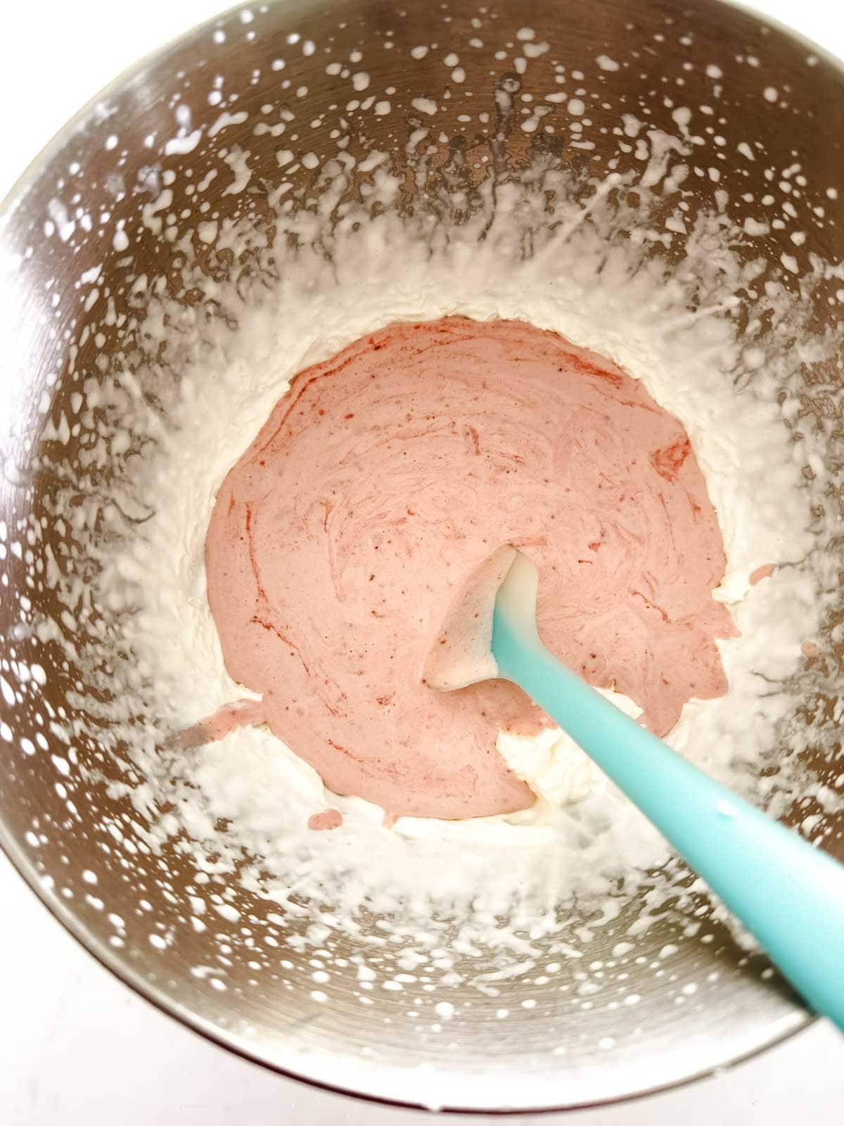 A metal mixing bowl with whipped cream and a pink strawberry mixture being gently folded together with a blue silicone spatula.