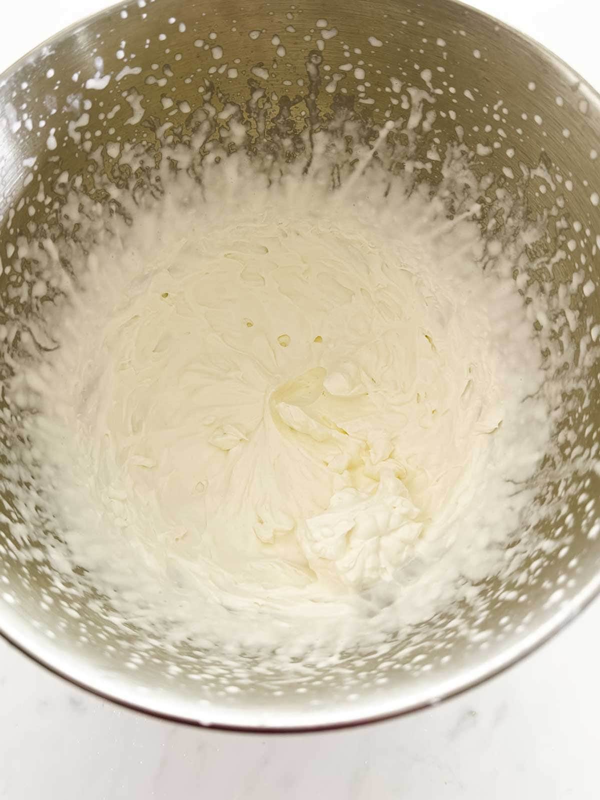 A metal mixing bowl containing freshly whipped cream, with some cream splattered on the sides of the bowl. The bowl sits on a light-colored surface.