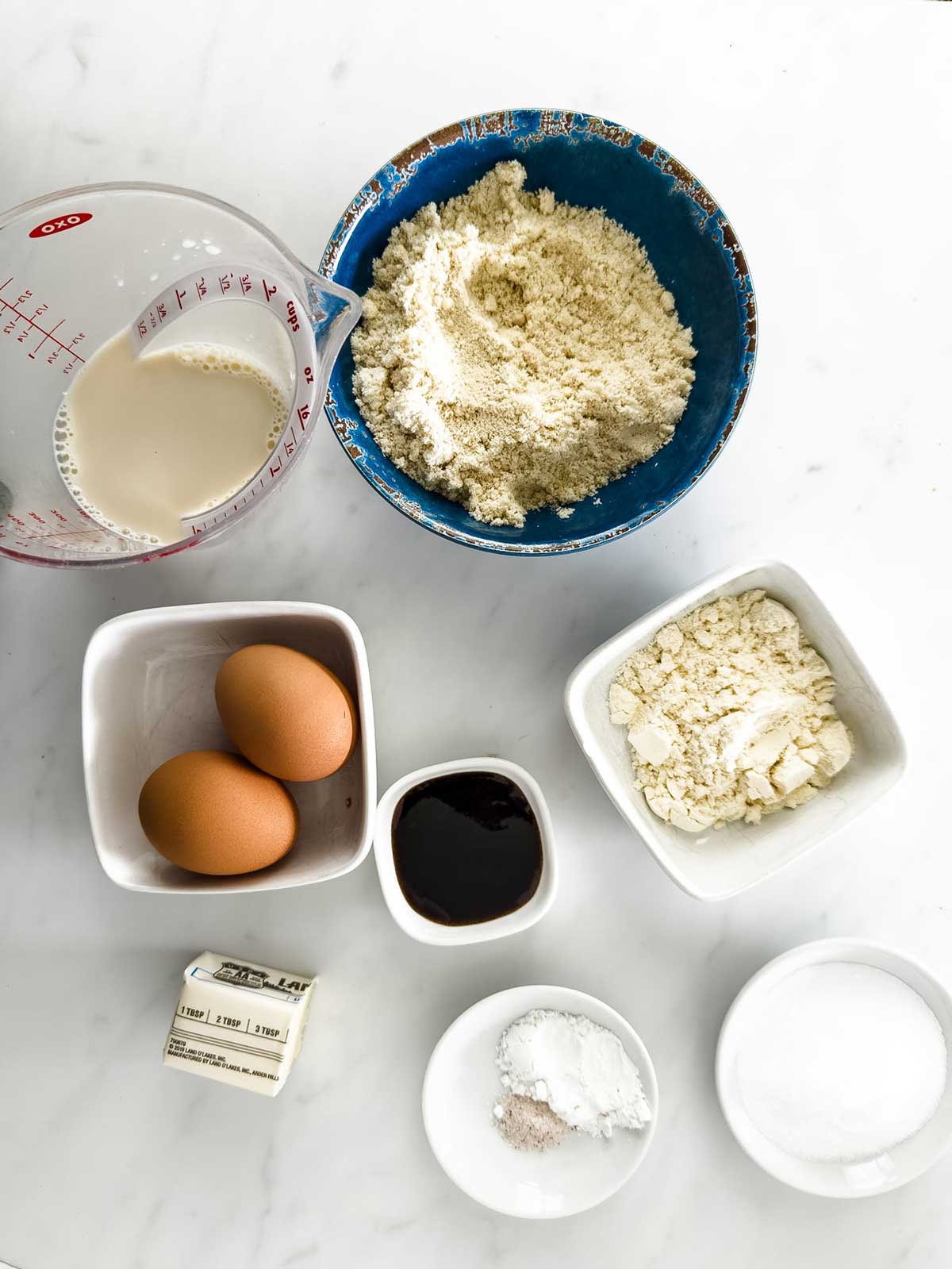 A flat lay of baking ingredients on a white surface: a blue bowl of almond flour, a measuring cup of milk, two eggs, butter, a small dish of vanilla, a bowl of flour, and small dishes of baking powder and sweetener.
