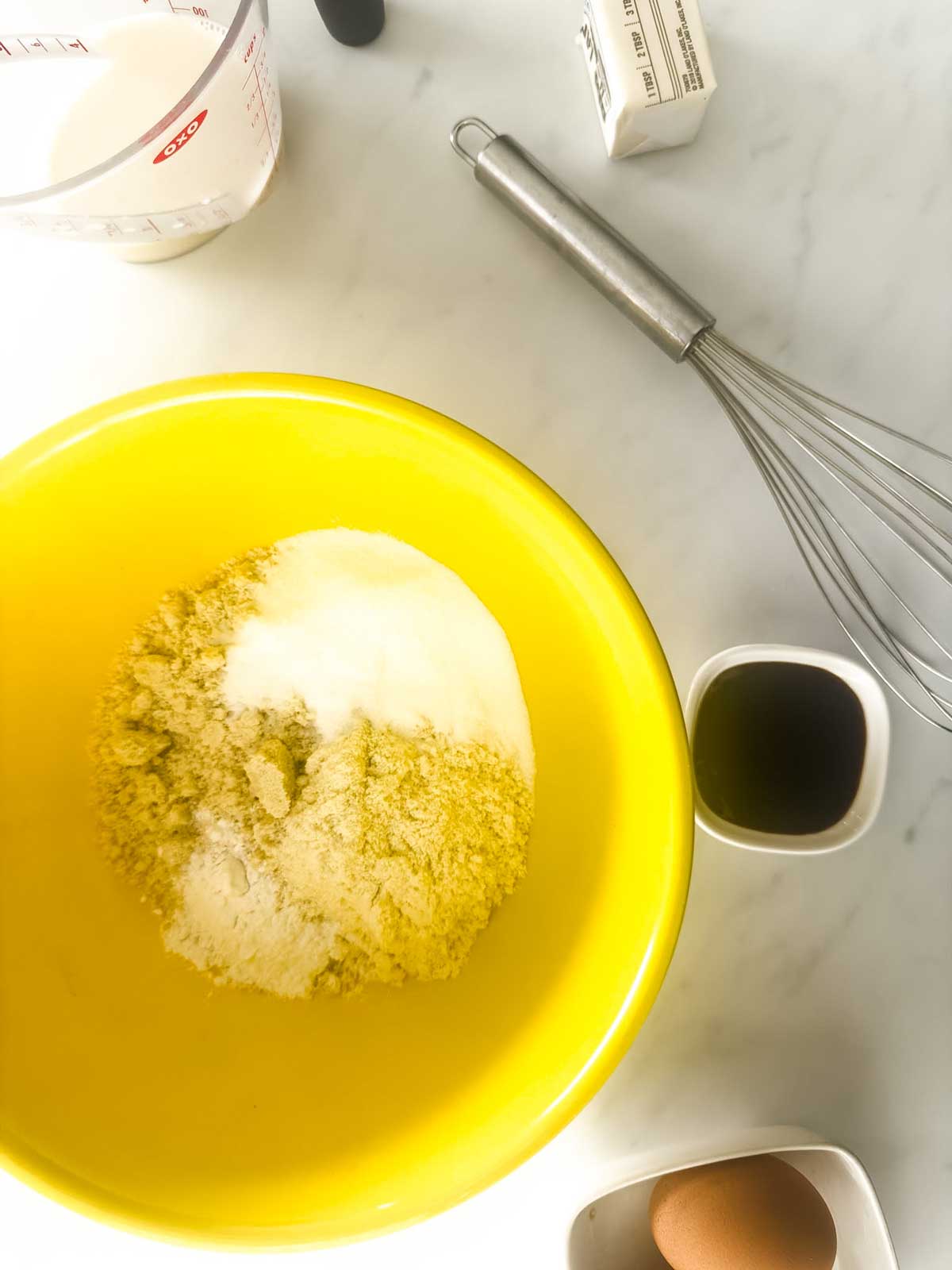 A yellow bowl with almond flour and sweetener sits on a white surface, surrounded by a whisk, measuring cup of milk, small bowl of vanilla extract, stick of butter, and a brown egg—everything you need to make delicious keto waffles.