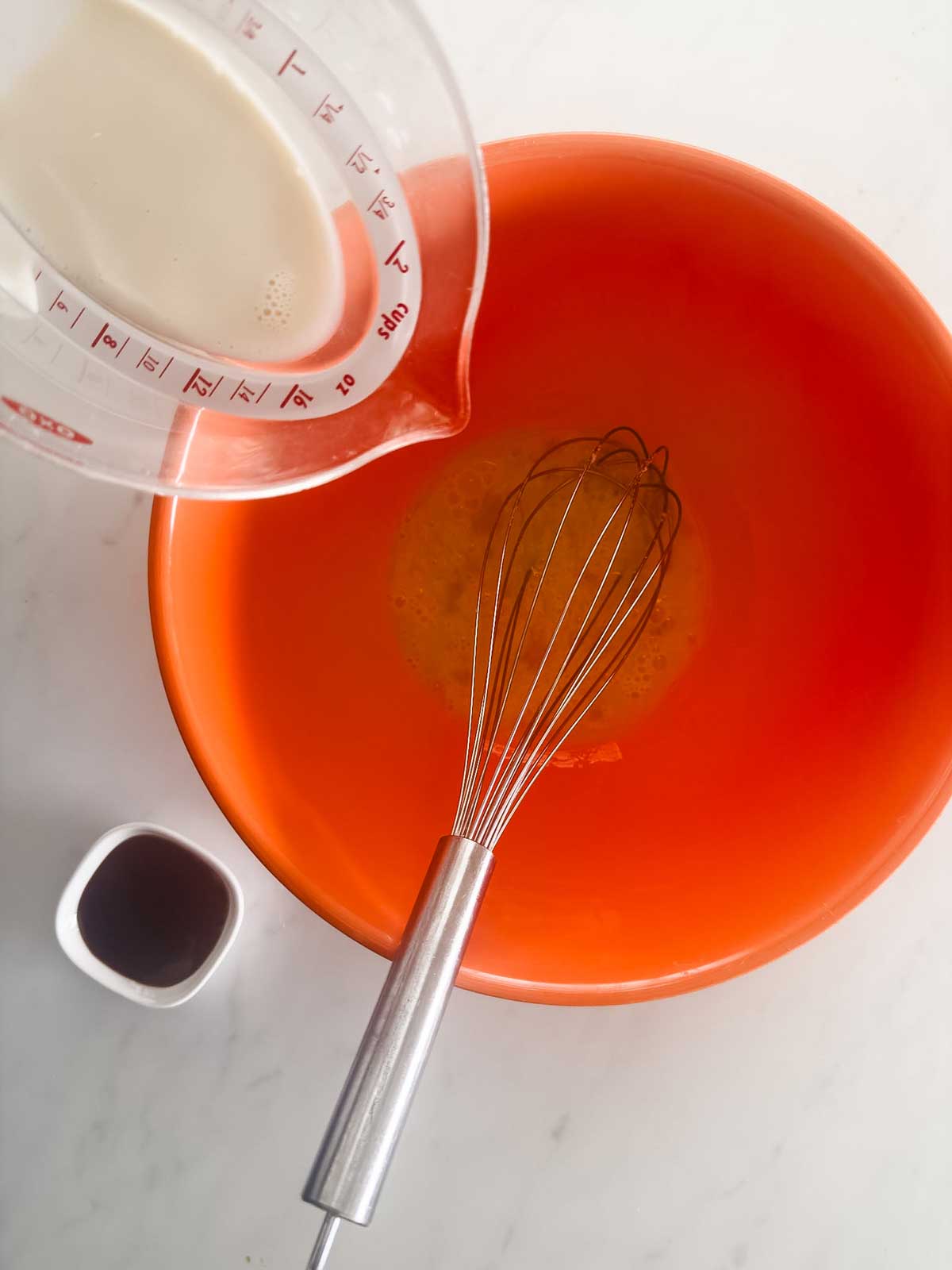 A measuring cup pours liquid into an orange mixing bowl containing a whisk and a yellow mixture. A small cup with a dark liquid sits next to the bowl on a white countertop.