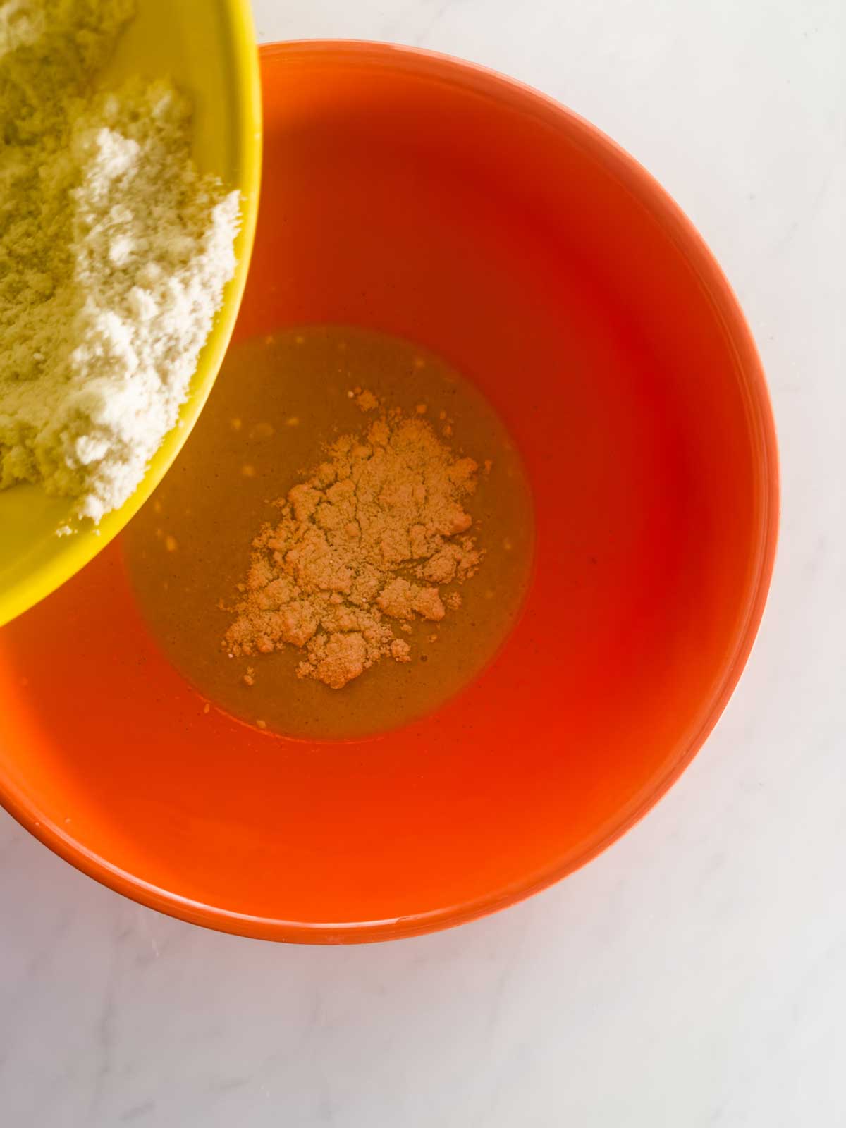 A yellow bowl pours flour into an orange bowl containing a liquid mixture, all placed on a white countertop.