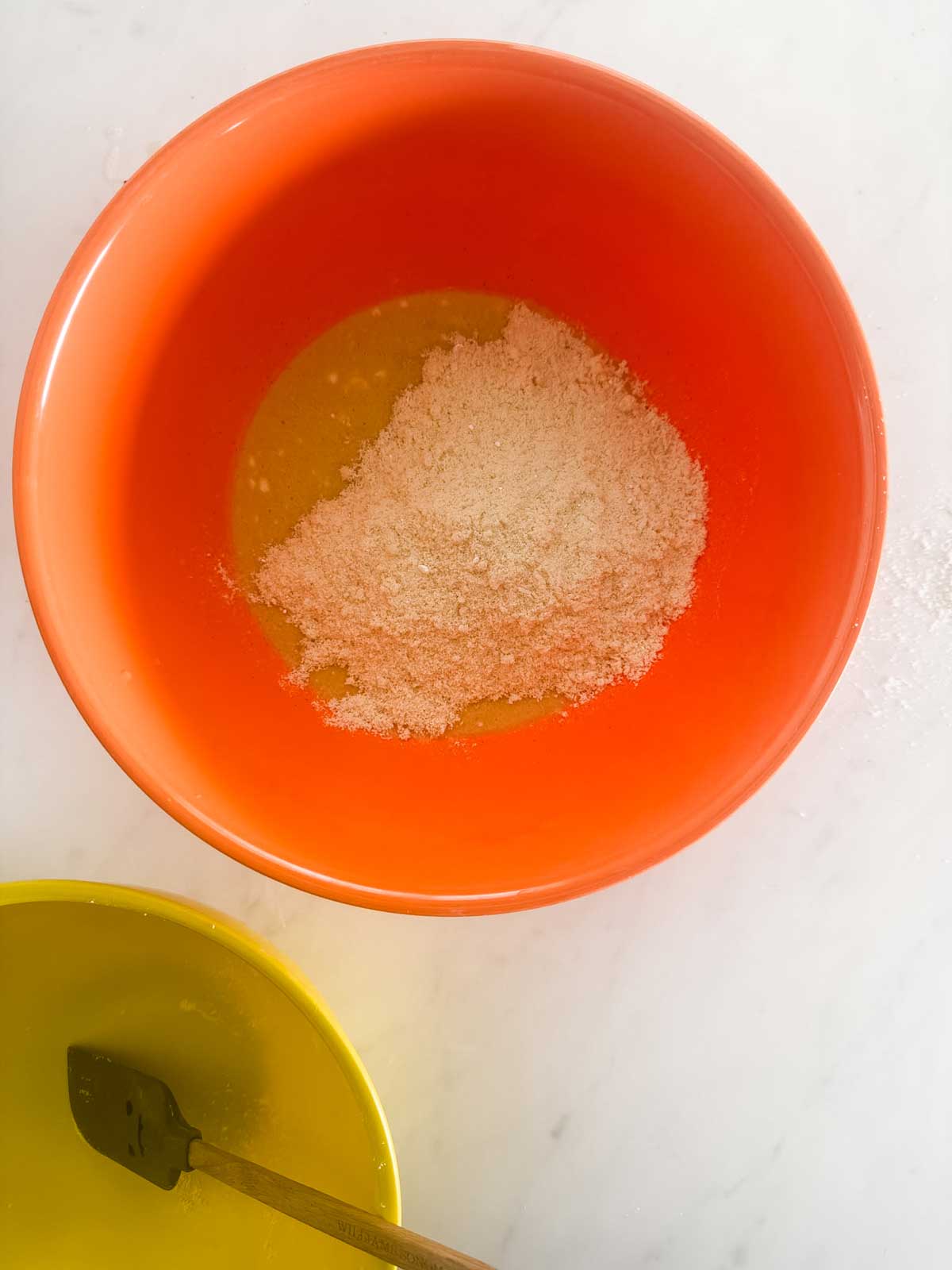 An orange mixing bowl with almond flour and liquid ingredients inside, next to a yellow bowl with a black spatula, all on a white countertop.