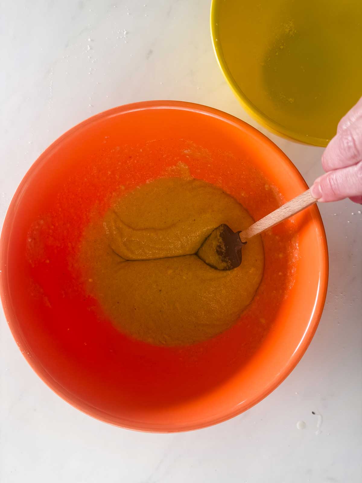 A person stirs a thick yellow-orange batter in an orange mixing bowl with a wooden spoon. A yellow bowl is partially visible nearby on a white countertop.