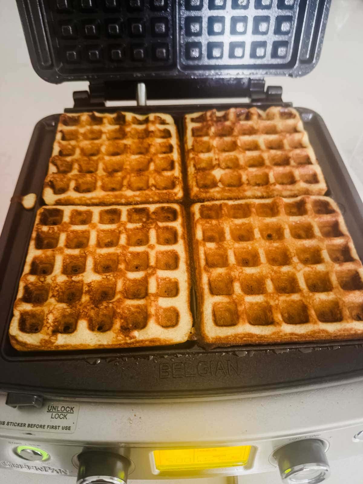 Four golden-brown waffles are cooking inside an open Belgian waffle maker. The waffles fill the four square sections of the waffle maker, which is sitting on a countertop.
