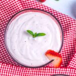 A glass of creamy strawberry mousse topped with a small mint sprig and a sliced strawberry on the rim, placed on a red and white checkered cloth.