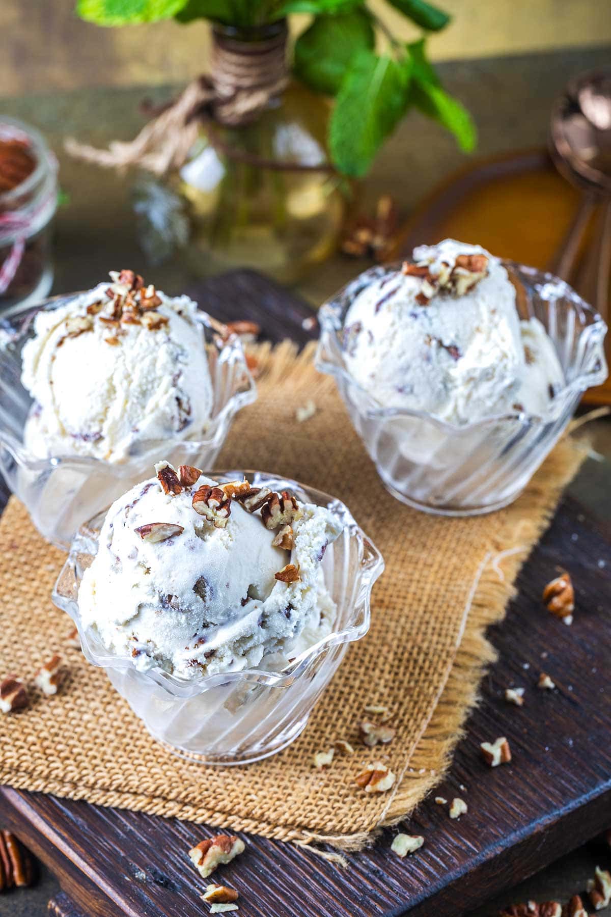 Three glass bowls filled with scoops of butter pecan ice cream mixed with chopped pecans sit on a piece of burlap atop a wooden board, with extra pecan pieces scattered around. A vase with green leaves is in the background.