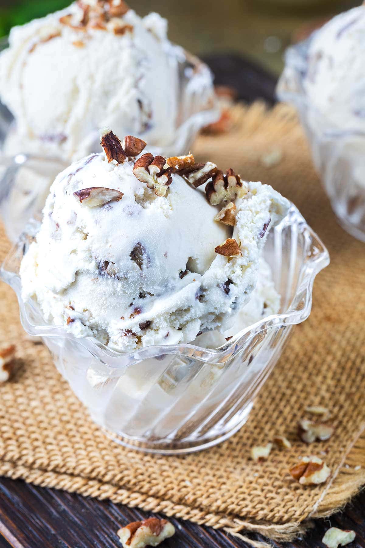 A glass bowl filled with scoops of creamy ice cream mixed with chopped pecans, topped with more pecans, sits on a woven mat with extra pecan pieces scattered around.