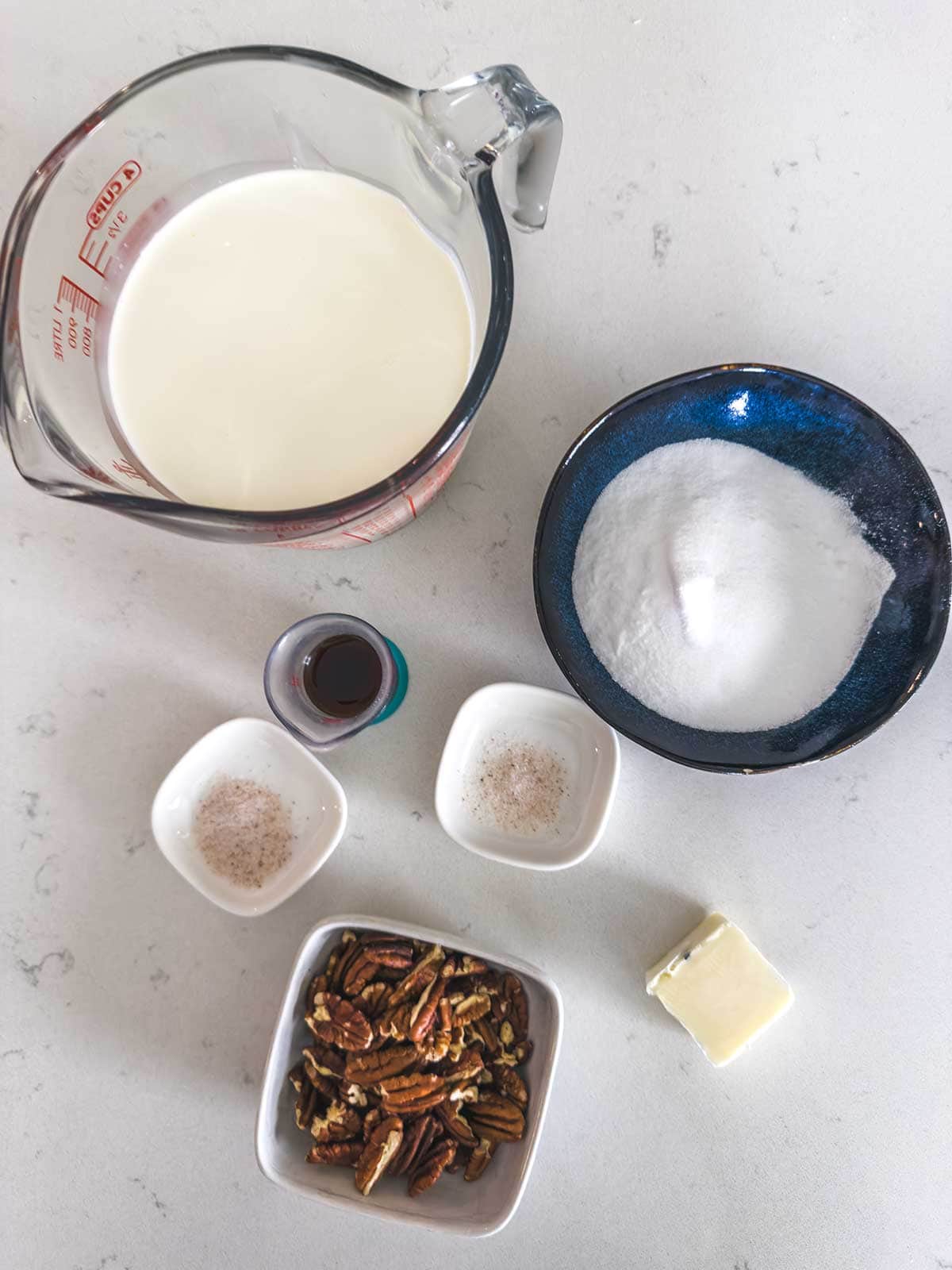 A top-down view of ingredients on a white surface: a measuring cup of milk, a bowl of sugar, a small bowl of pecans, a pat of butter, a small bowl of vanilla extract, and two small dishes with spices.