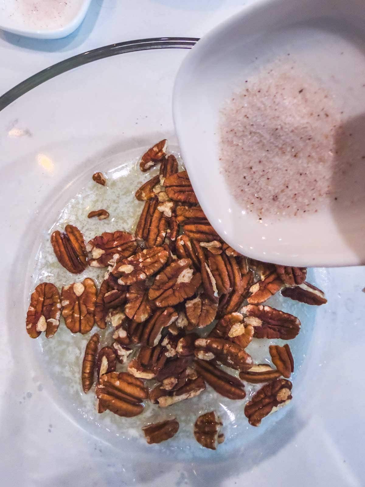 Pecans in a glass bowl are being topped with a white, creamy liquid that has specks of spice, likely being poured from a small white dish.