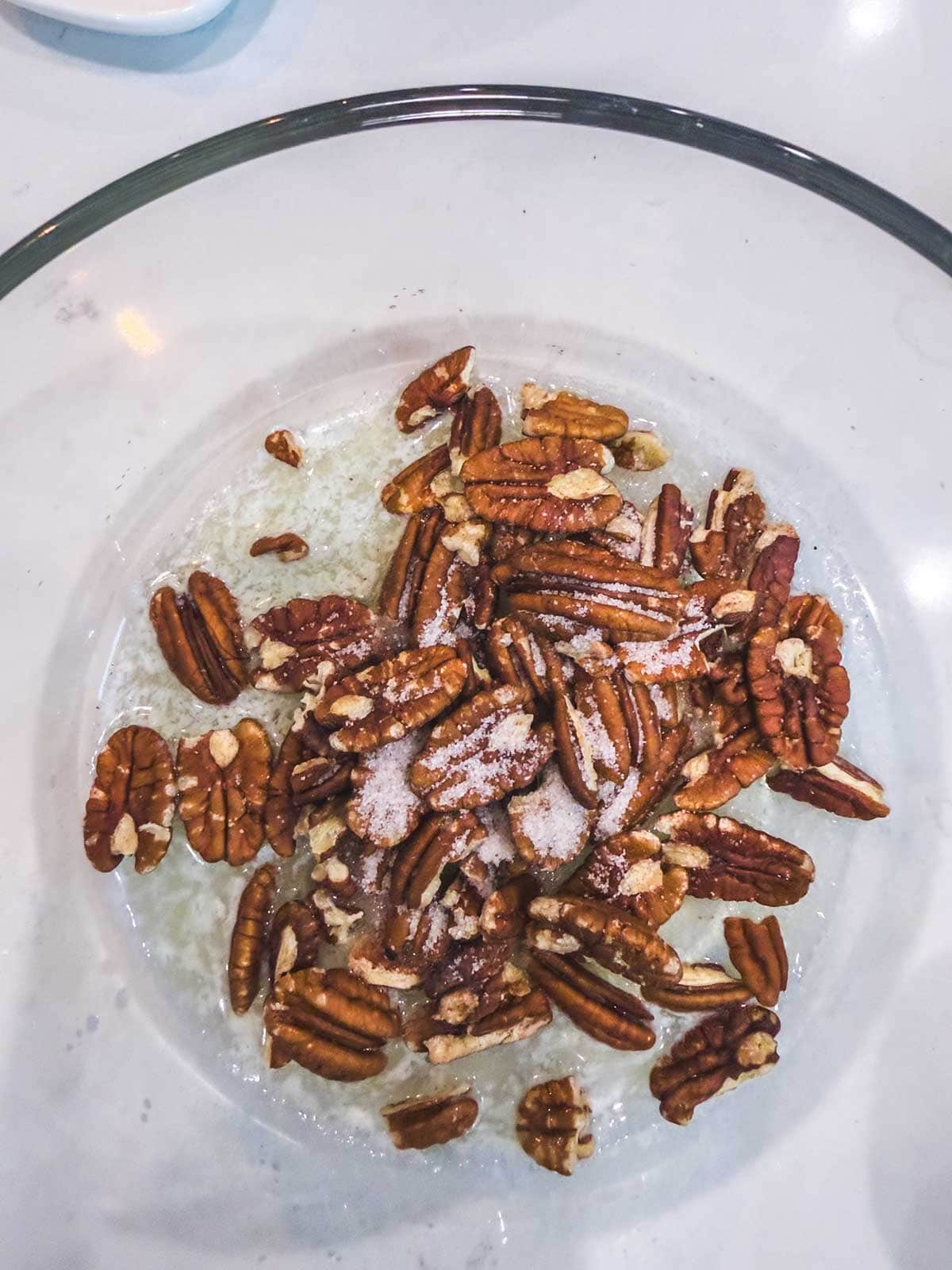 Pecan halves and granulated sugar in a clear glass bowl with melted butter on the bottom, set on a white countertop.