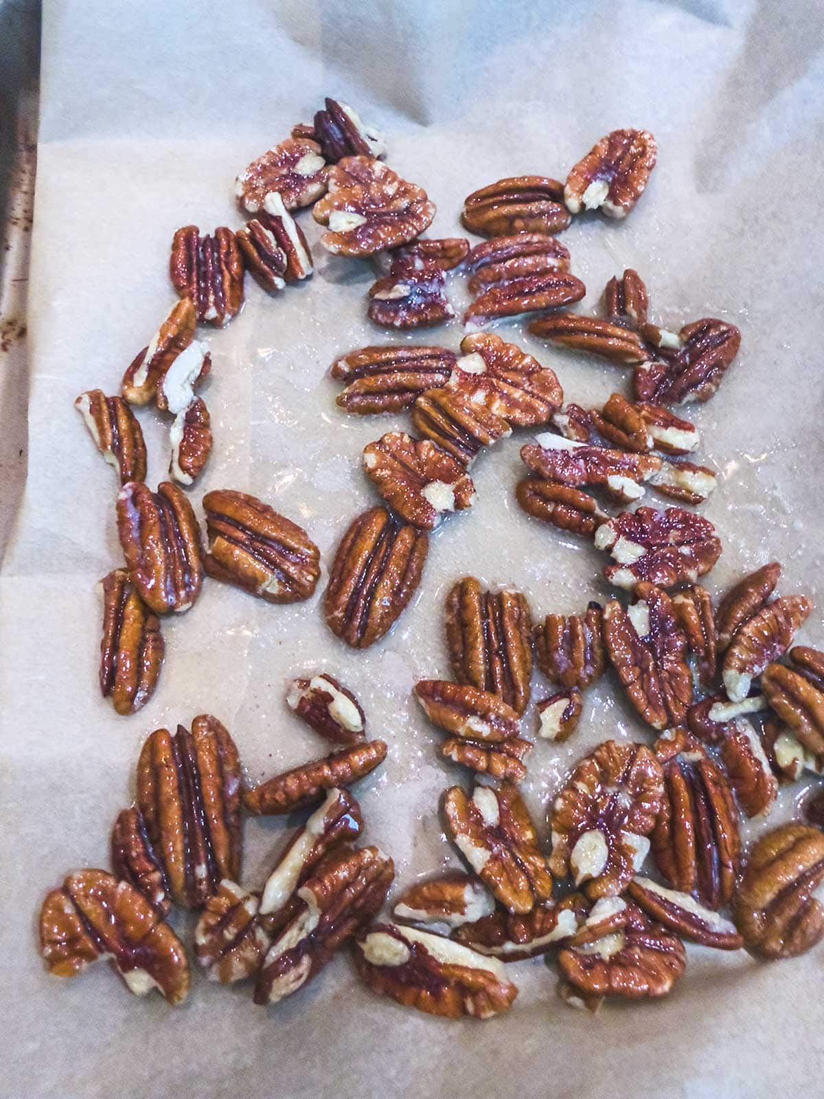 Pecan halves coated in a shiny glaze are spread out on a sheet of parchment paper, likely for cooling after roasting or candying.