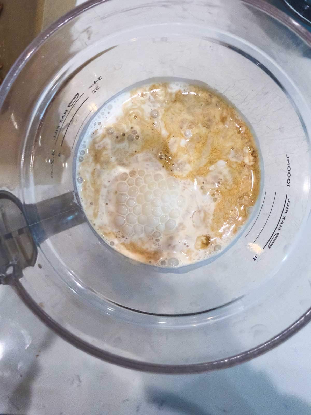 A close-up view of a mixing bowl containing a frothy mixture of yeast, water, and flour, seen from above. Measurement markings are visible on the side of the transparent bowl.