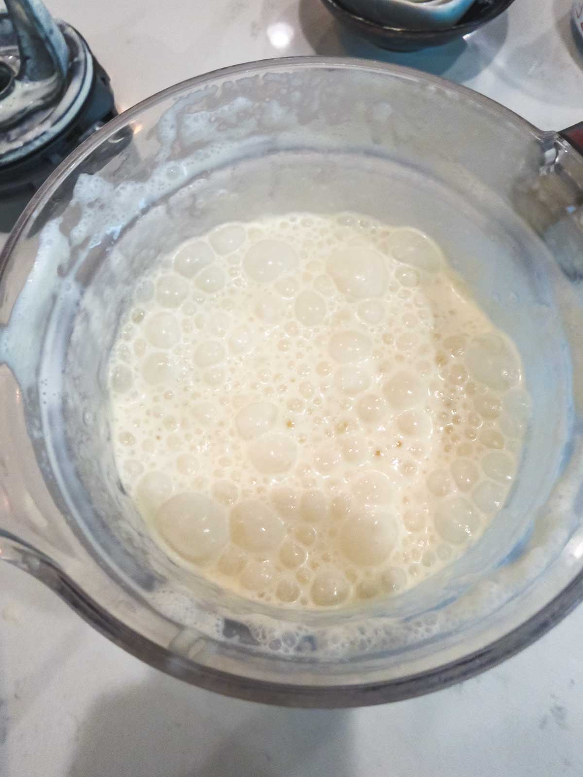 A clear blender pitcher filled with frothy, bubbly pale liquid sits on a white countertop, with part of a blender base visible nearby.