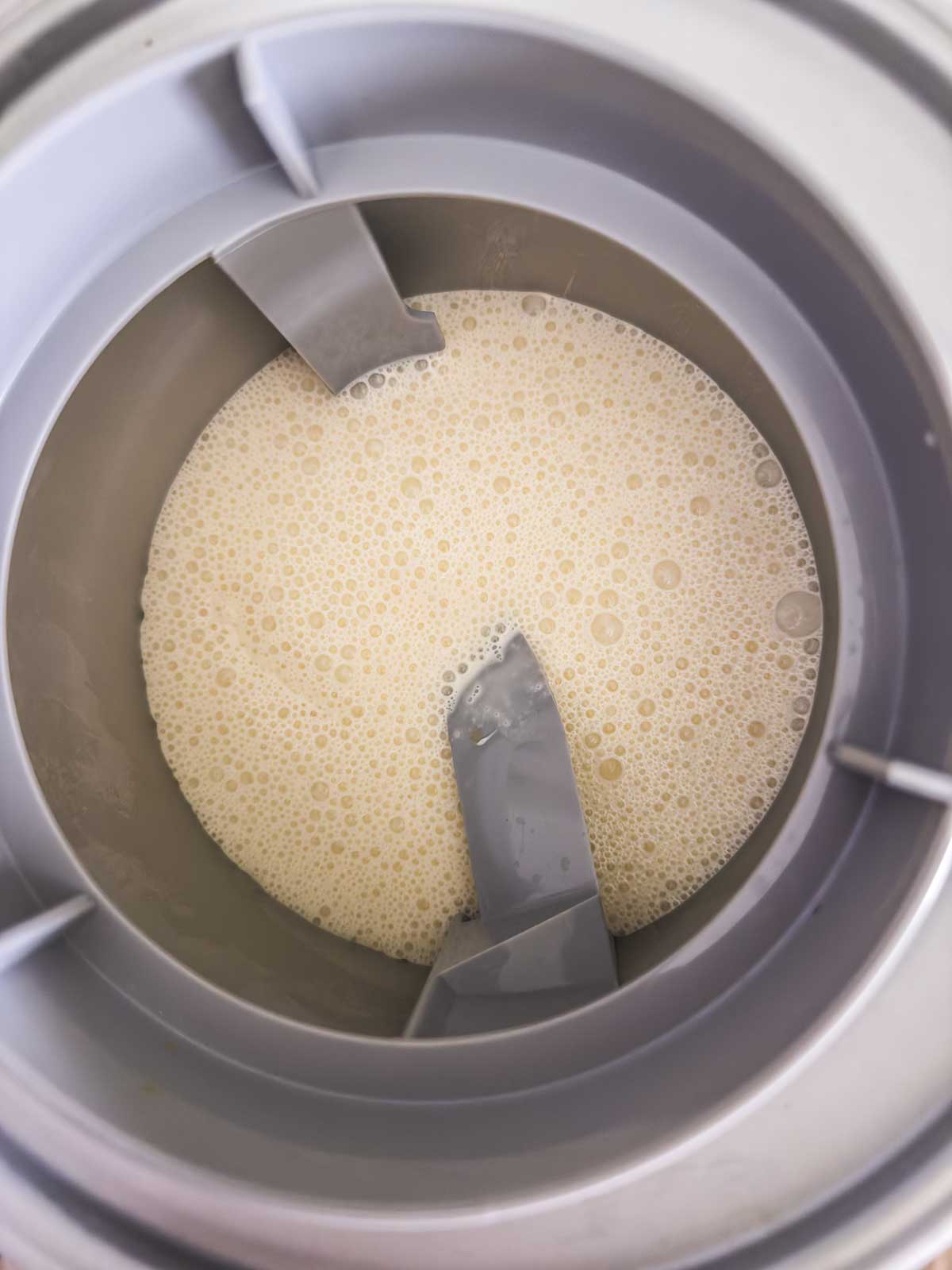 A top-down view of a metal ice cream maker bowl filled with a light beige, frothy liquid mixture, with two gray blades in the center.