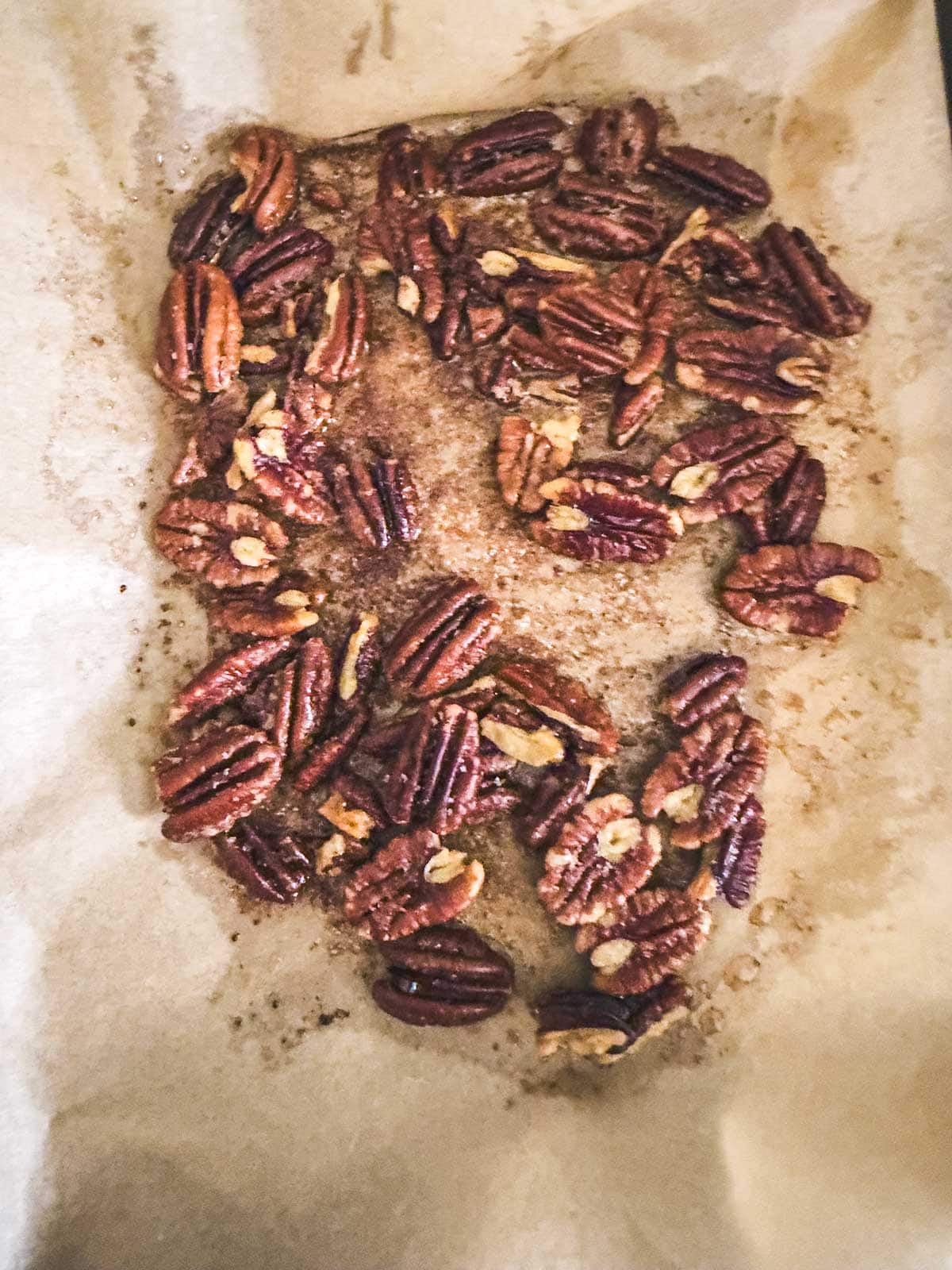 Toasted pecans spread out on a sheet of parchment paper, showing a slightly browned color and glistening coating, likely from roasting or seasoning.