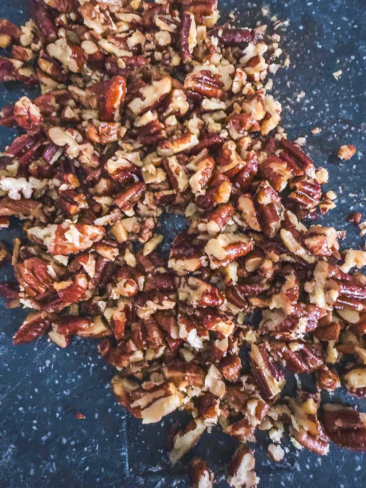 Chopped pecans spread across a dark, speckled surface. The nuts are unevenly cut, showing a mix of small and medium pieces with brown shells and lighter interiors.