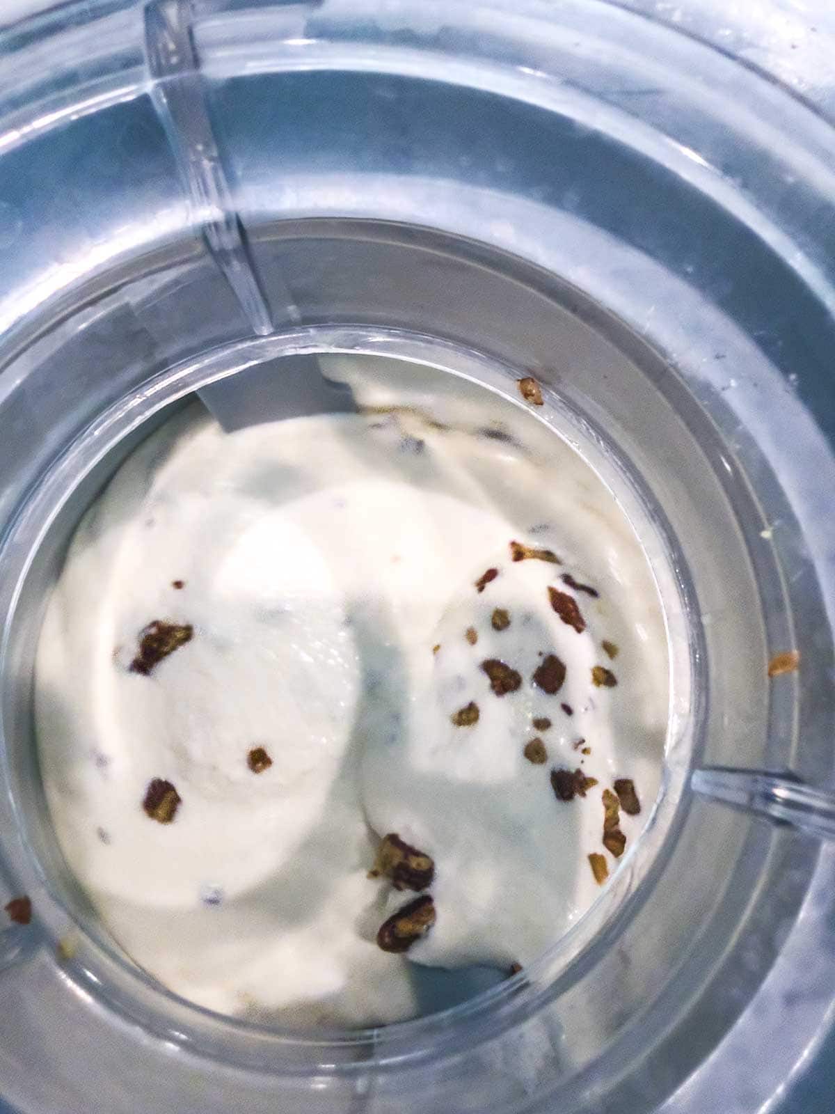 Close-up view of creamy ice cream being churned in an ice cream maker, with small chocolate or nut pieces mixed into the vanilla mixture.