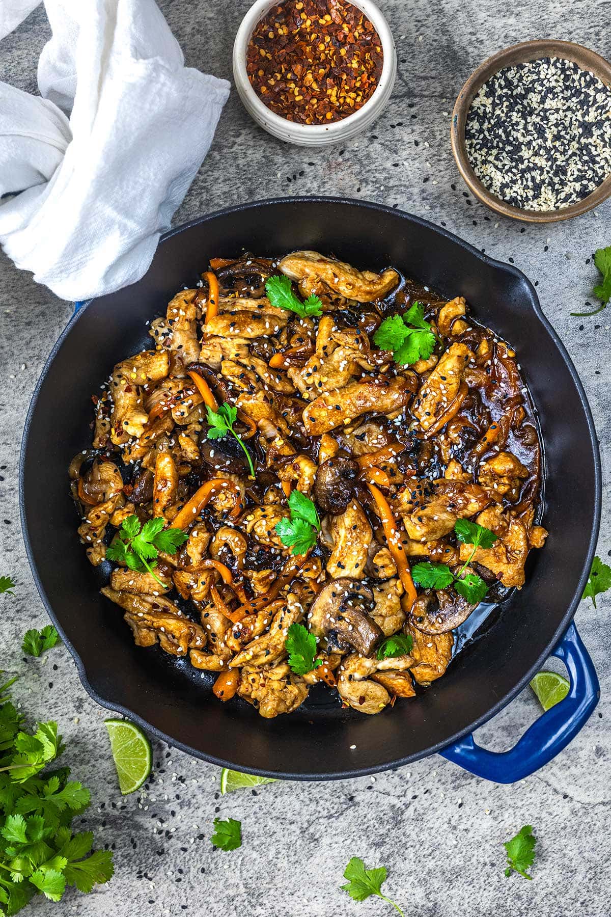 A skillet filled with stir-fried chicken, mushrooms, and carrots in a glossy sauce, garnished with sesame seeds and cilantro. Bowls of red pepper flakes and sesame seeds, lime wedges, and fresh herbs are nearby.