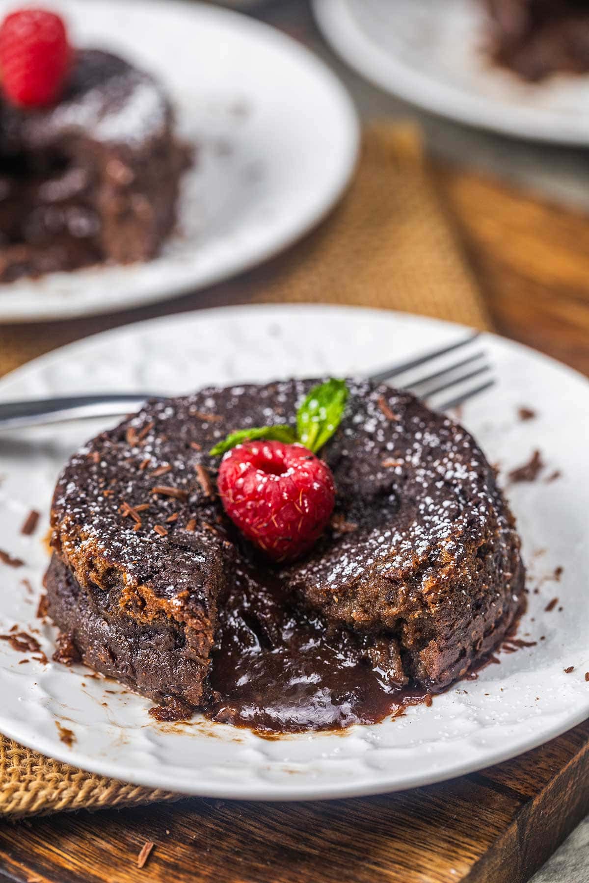 A chocolate lava cake with a molten center sits on a white plate, garnished with a raspberry, mint leaves, and powdered sugar. A fork rests beside the dessert, and a piece is cut out, showing the gooey chocolate filling.