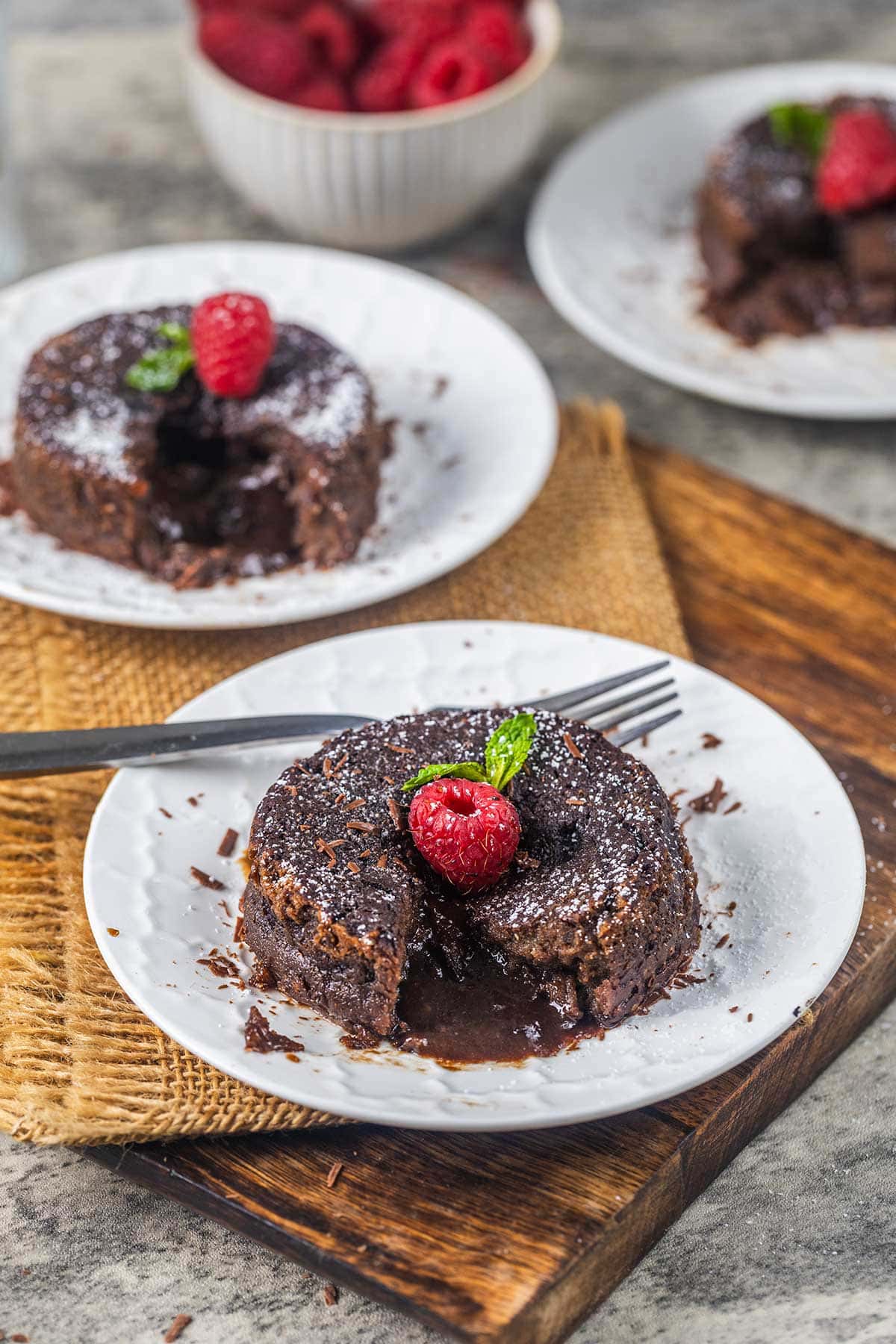 A chocolate lava cake topped with a raspberry and mint leaf sits on a white plate, with a fork and a bowl of raspberries in the background. Another plate with cake is visible behind it.
