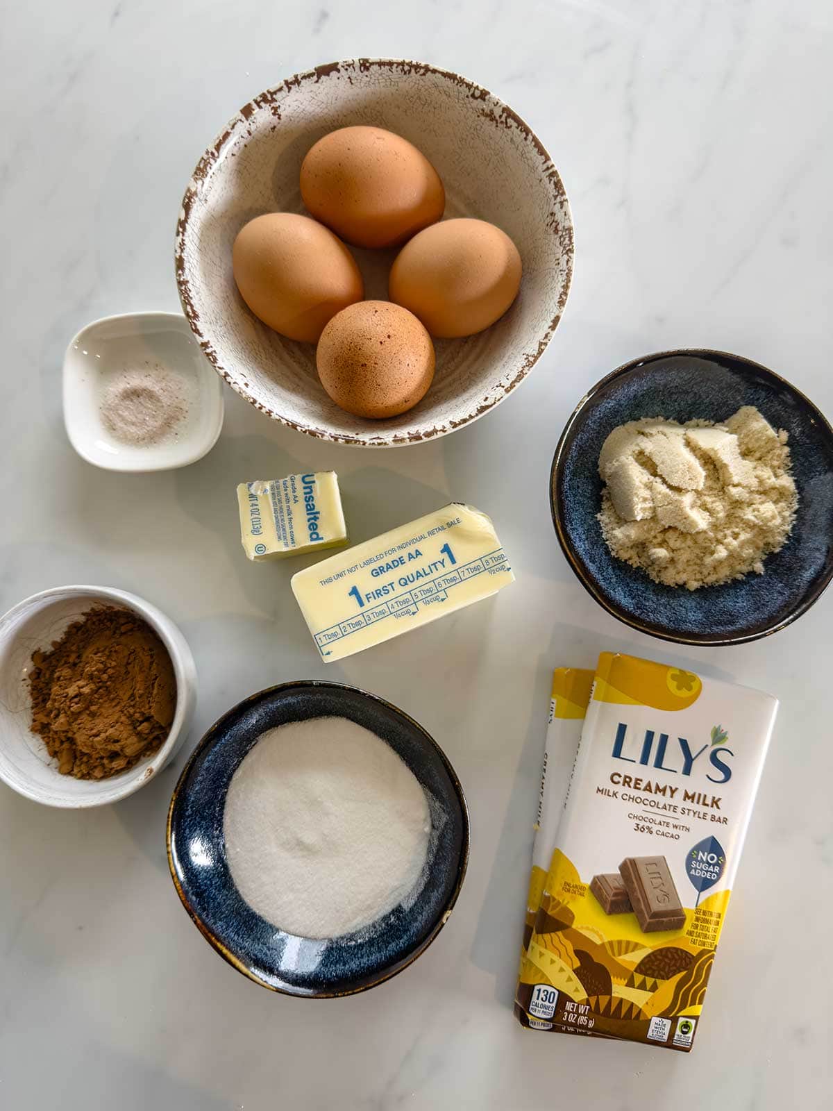 A top view of baking ingredients on a white surface, including three eggs in a bowl, two butter sticks, a bowl of almond flour, a bowl of sugar, a bowl of cocoa powder, a small dish of salt, and two Lilys milk chocolate bars.