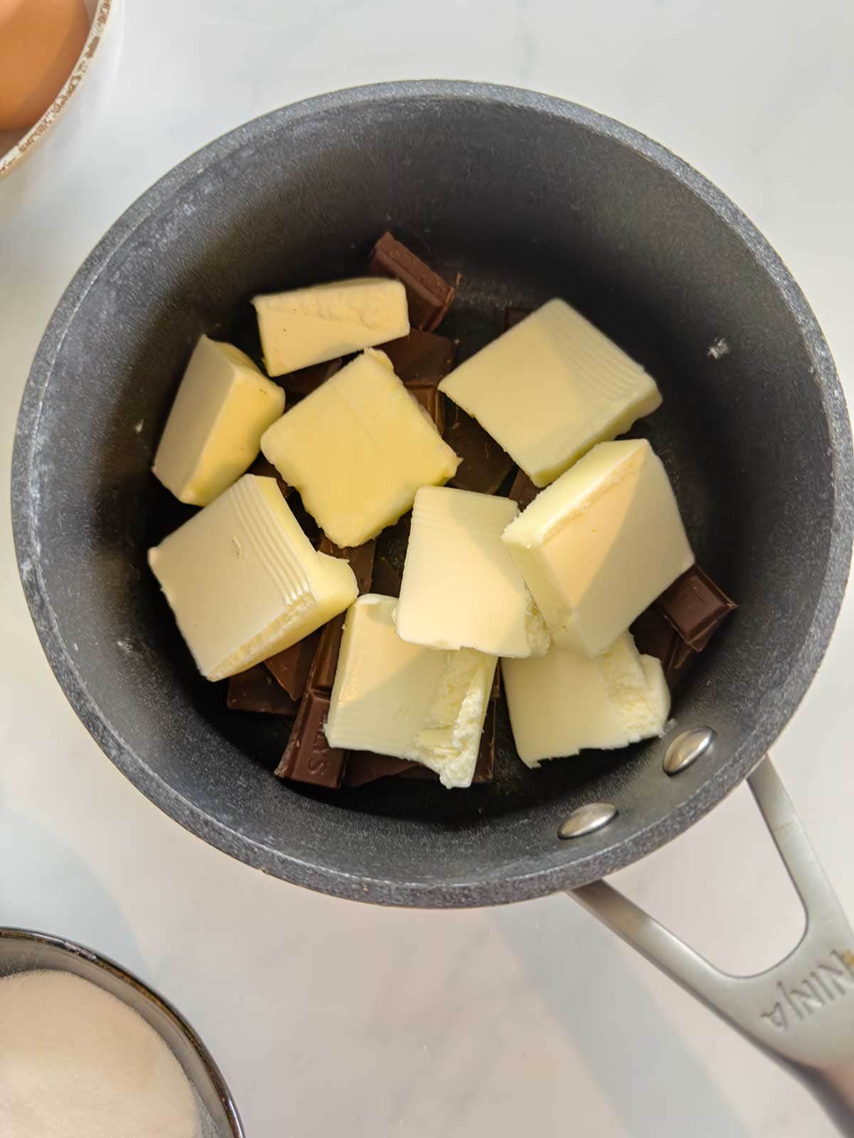 A saucepan containing chunks of butter and pieces of chocolate, ready to be melted, sits on a white countertop. A bowl with sweetener and part of an egg are visible nearby.