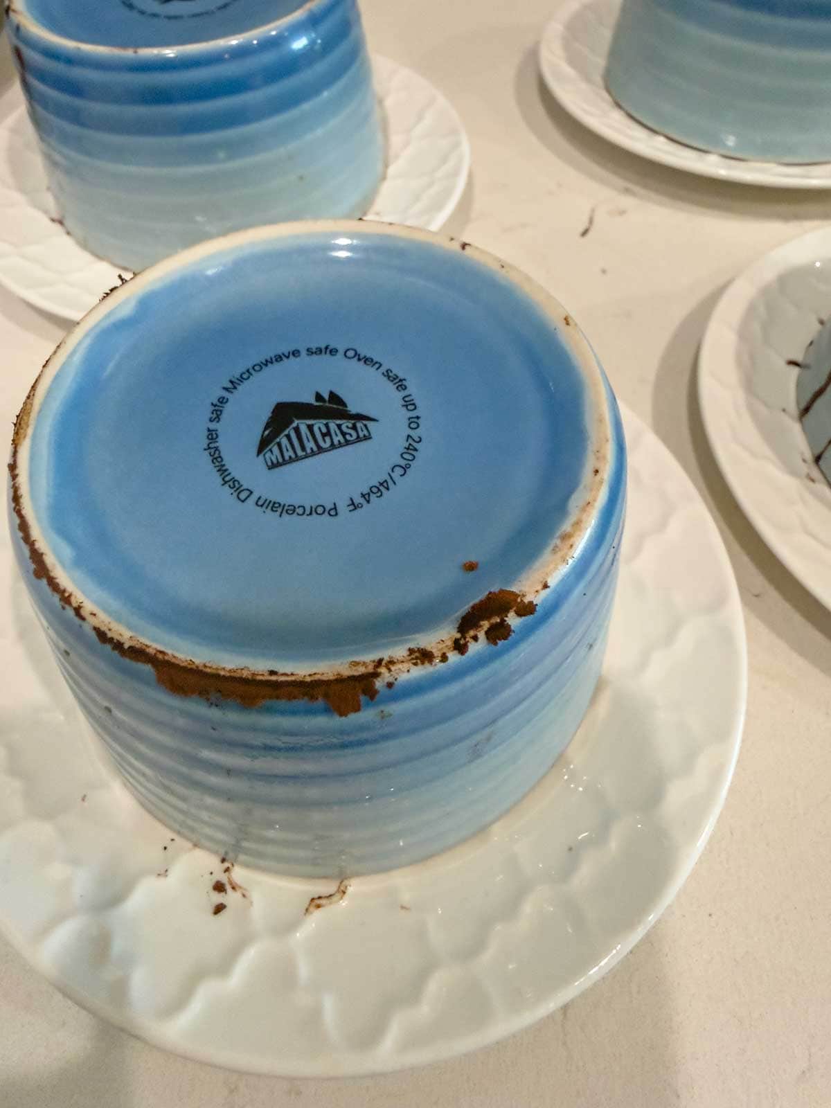 A blue ceramic bowl with brown stains on its base is placed upside down on a white plate. The bowl is labeled “Malacasa” and “Microwave safe, Oven safe, Porcelain Dishwasher safe.” Other plates are partially visible.