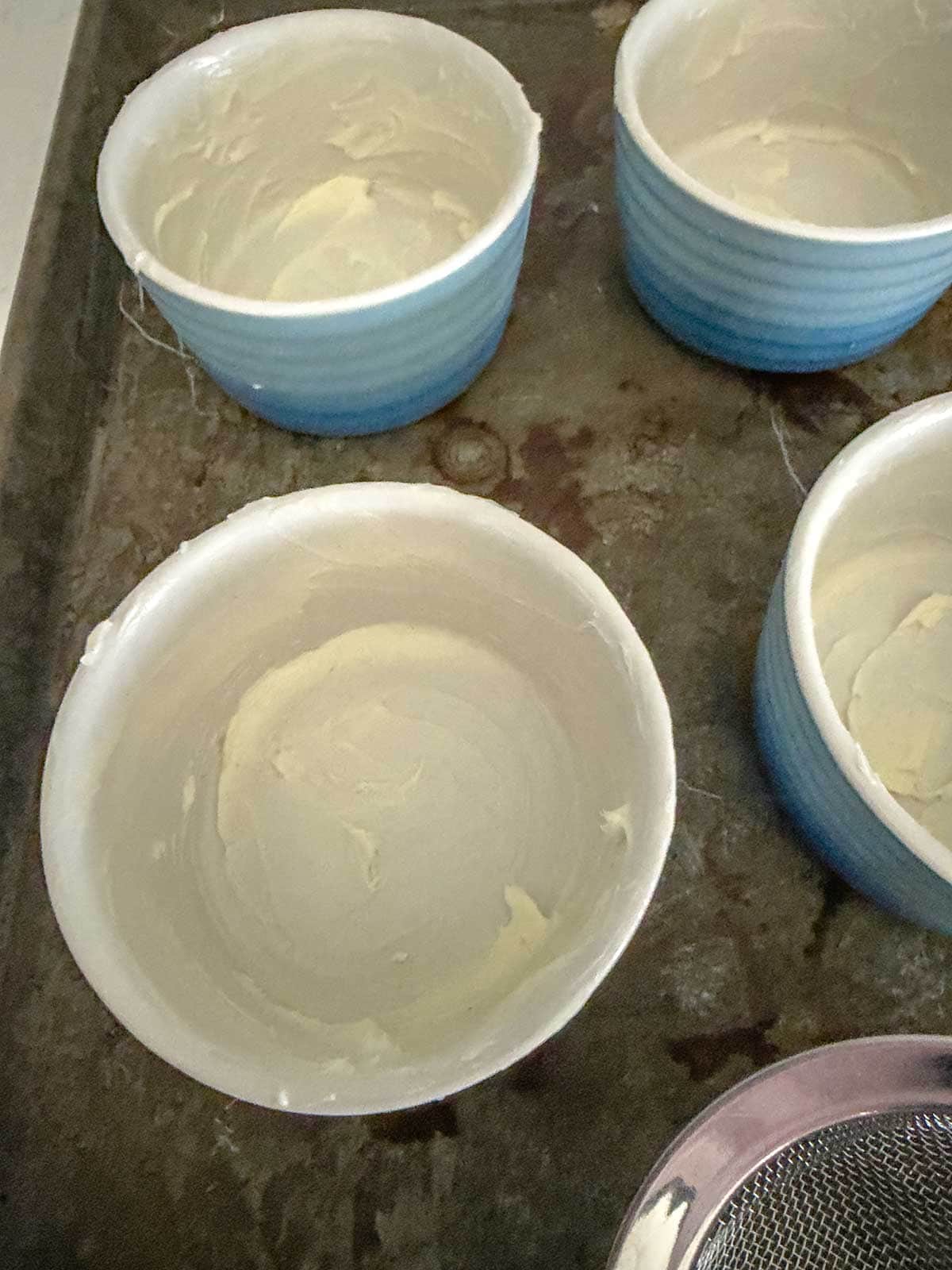 Four white and blue ramekins coated with butter are placed on a dark metal baking sheet, ready for baking preparation.