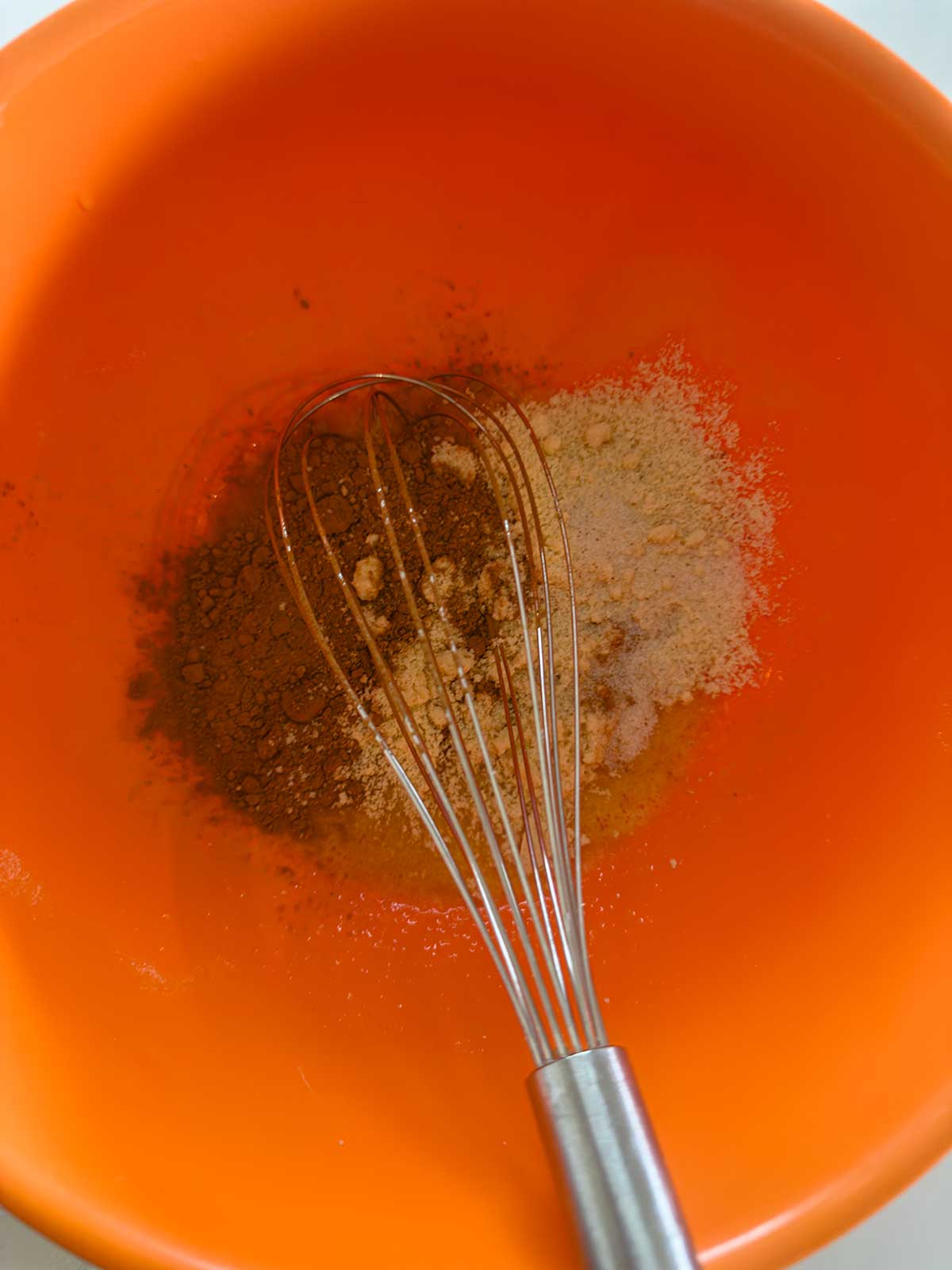 A metal whisk in an orange bowl with piles of brown and tan powdered spices, ready to be mixed.