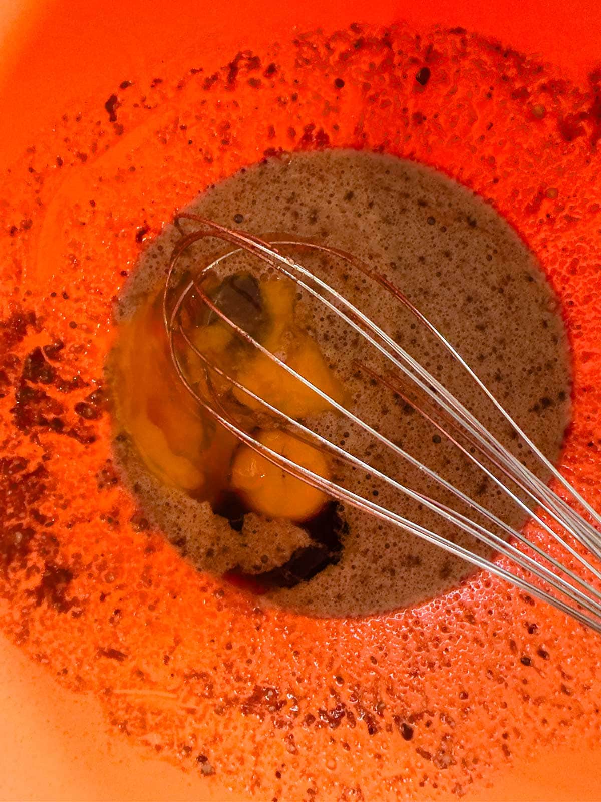 A close-up of a metal whisk mixing eggs, sweetener, and cocoa powder in an orange bowl, with the ingredients partially combined.