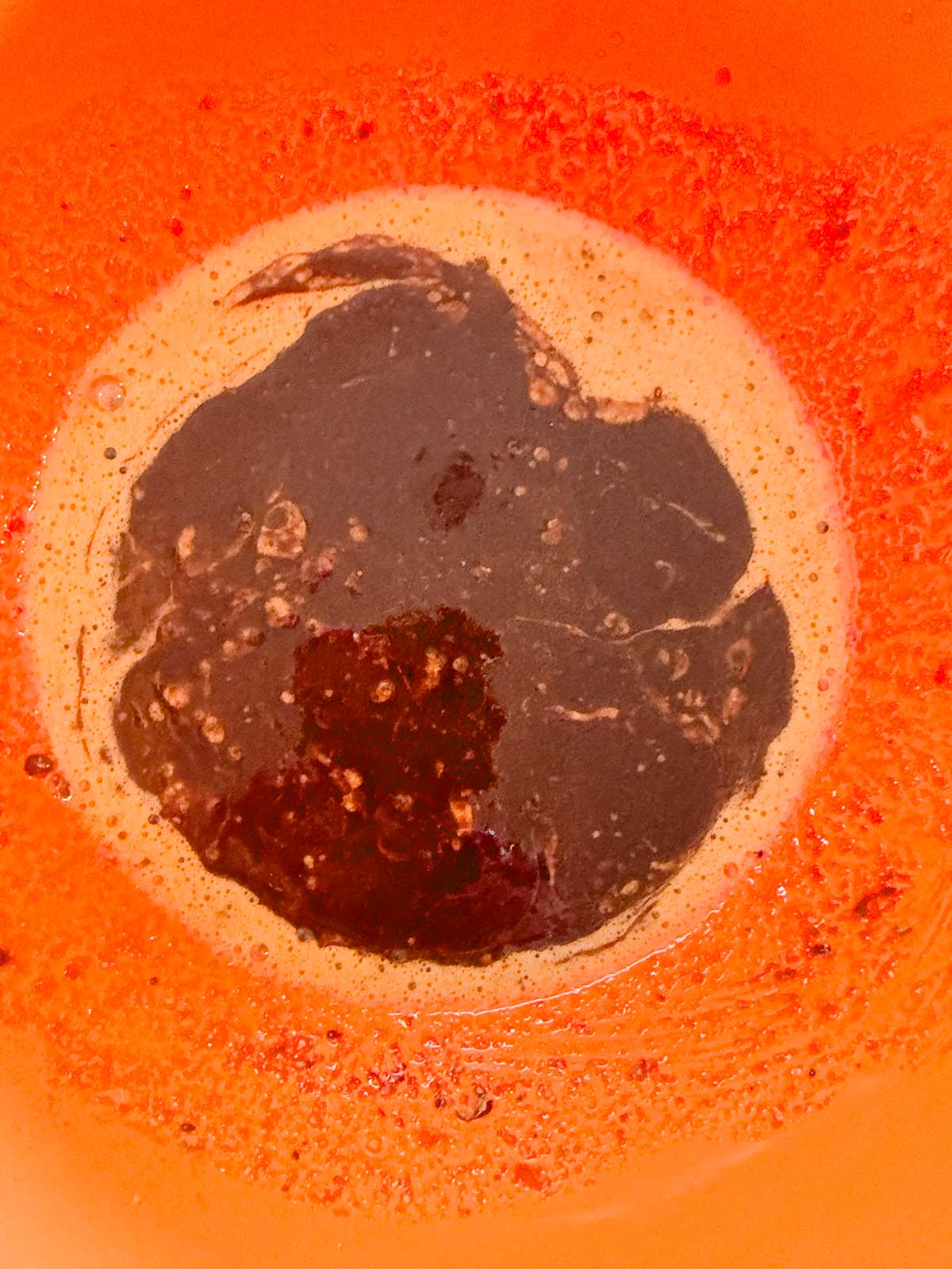 A close-up of a bright orange bowl with a dark brown, thick liquid pooled in the center, surrounded by frothy bubbles and some lighter brown residue along the sides.