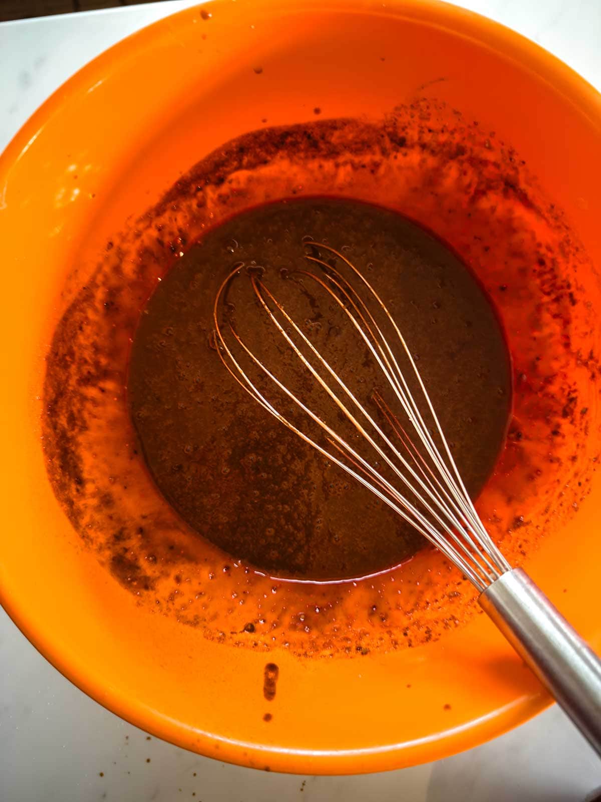 A metal whisk rests in an orange mixing bowl containing a dark brown liquid mixture, possibly for baking, with some residue on the sides of the bowl.