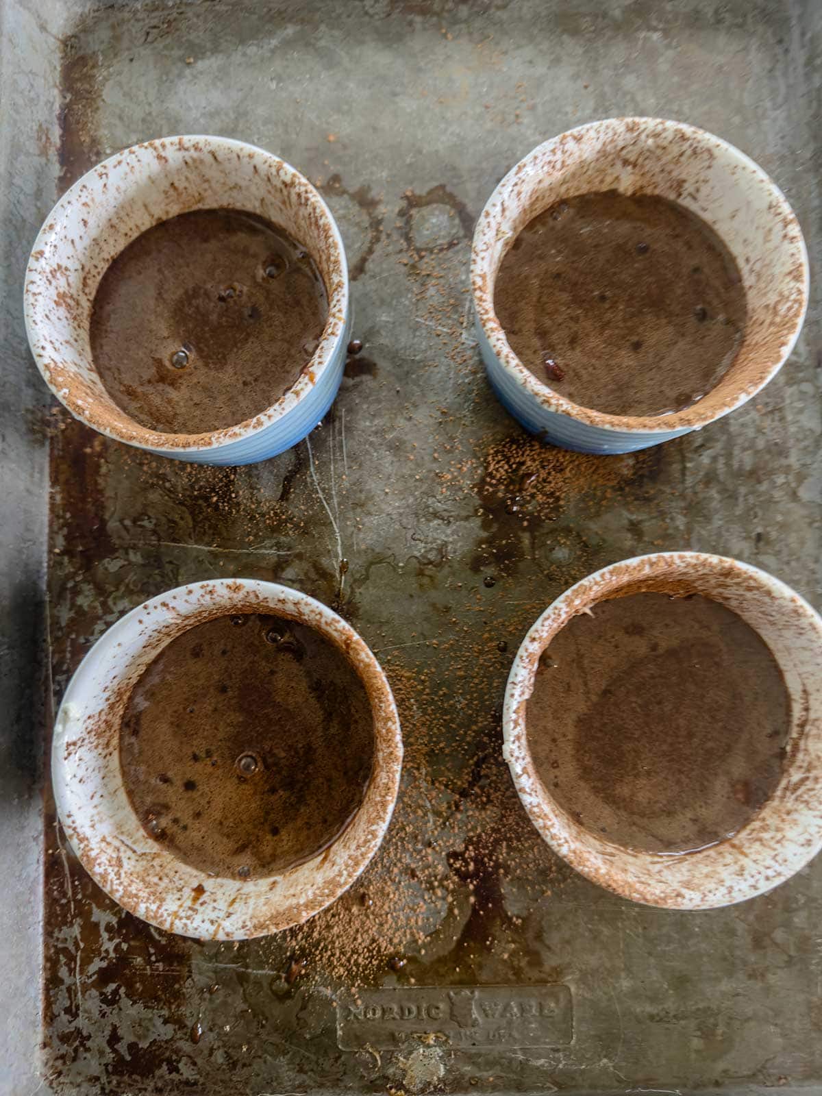 Four white ramekins filled with chocolate batter are placed on a metal baking tray. The rims of the ramekins are dusted with cocoa powder and the tray has splashes of batter and cocoa.