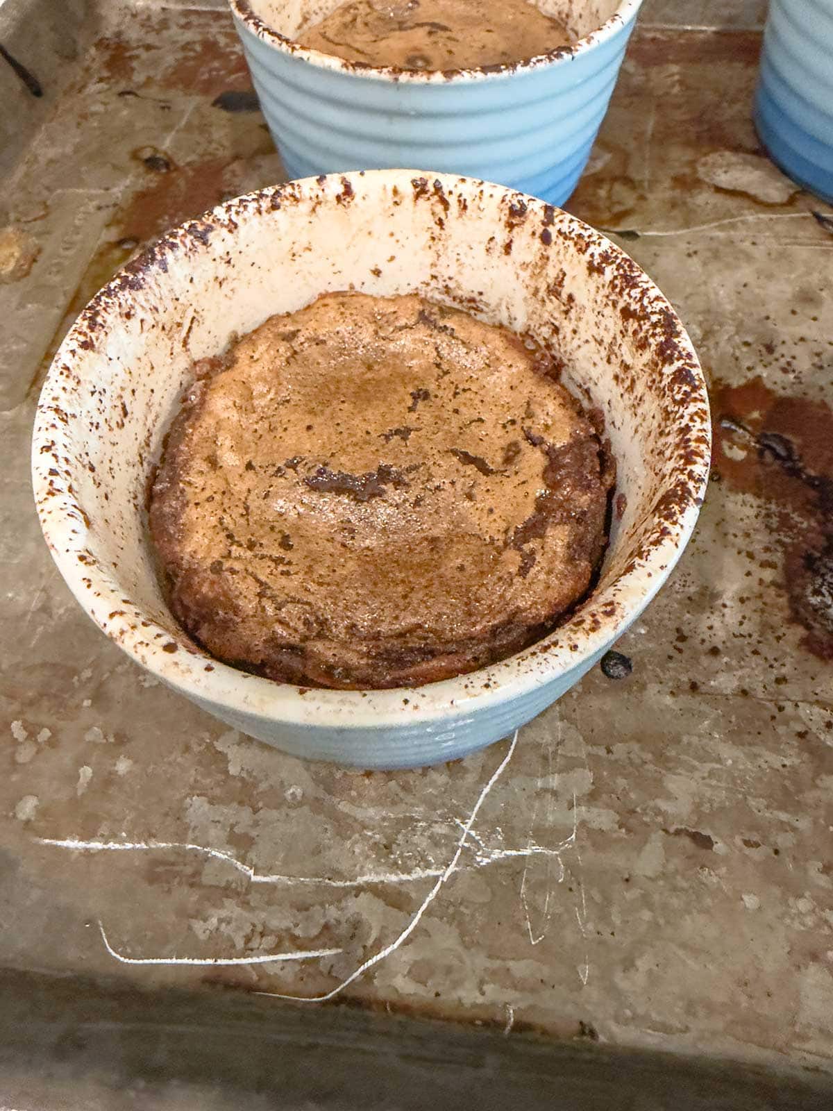 A blue ramekin filled with a baked chocolate dessert, possibly a souffle or lava cake, sits on a stained and well-used metal baking sheet. Another ramekin is partially visible in the background.