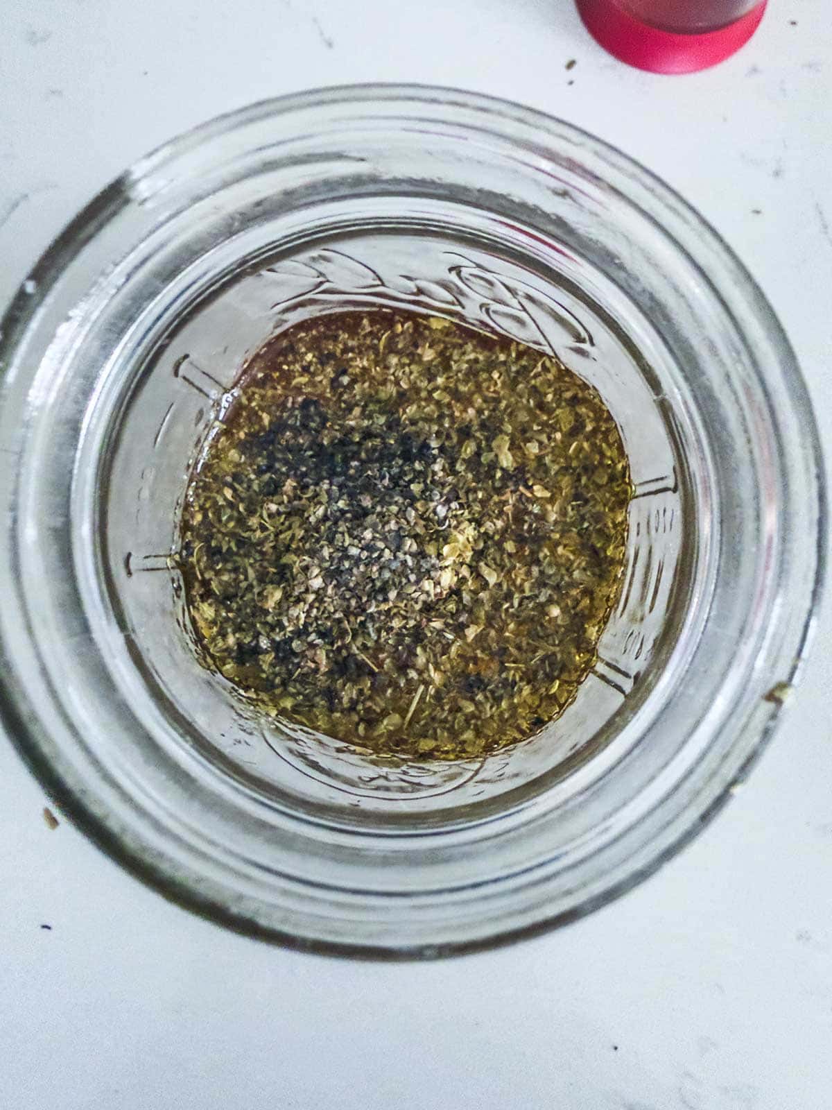 A glass jar seen from above contains a mixture of olive oil, dried herbs, and ground black pepper on a white surface.