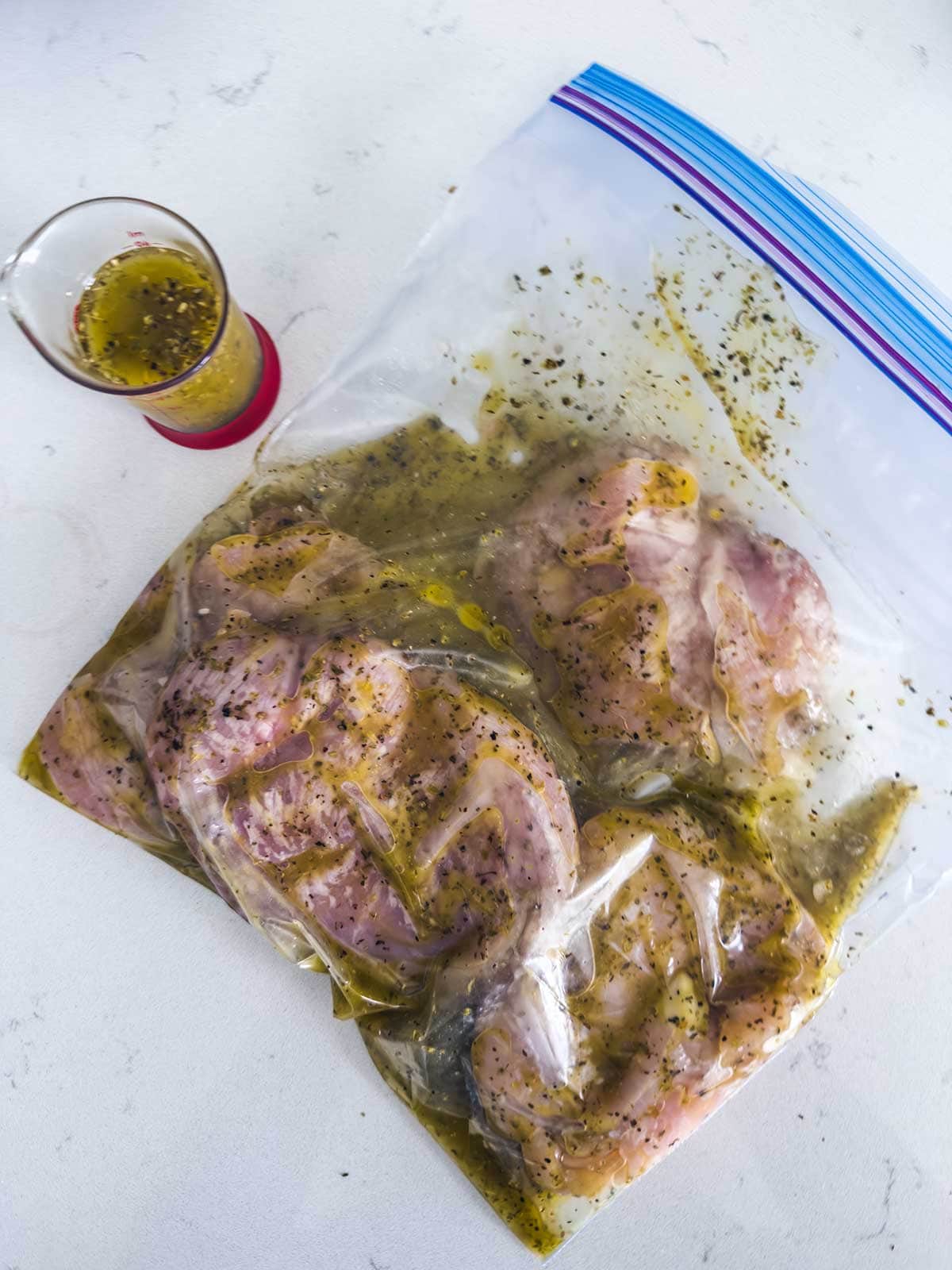 A plastic zip-top bag filled with raw chicken breasts marinating in a herb and oil mixture sits on a white countertop next to a small glass measuring cup containing leftover marinade.