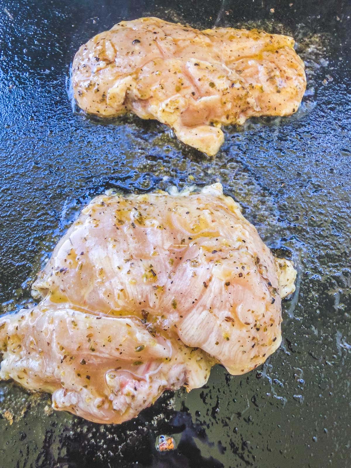 Two pieces of seasoned raw chicken breast cooking on a black griddle, with visible oil and spices on the meat and cooking surface.