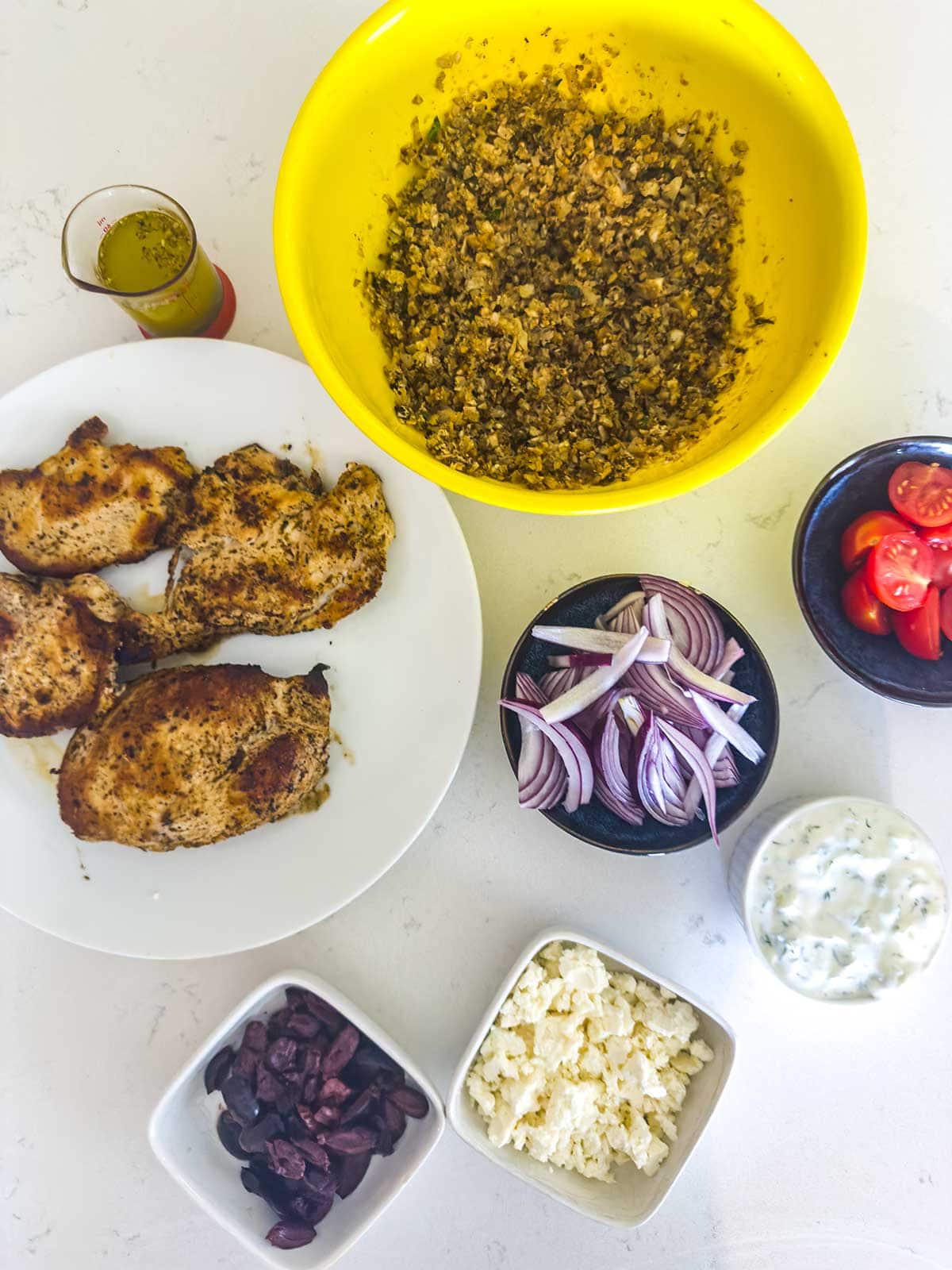Top-down view of ingredients for a Mediterranean meal: grilled chicken breast, chopped olives, crumbled feta, sliced red onions, grape tomatoes, tzatziki sauce, olive oil, and a bowl with a herb mixture.