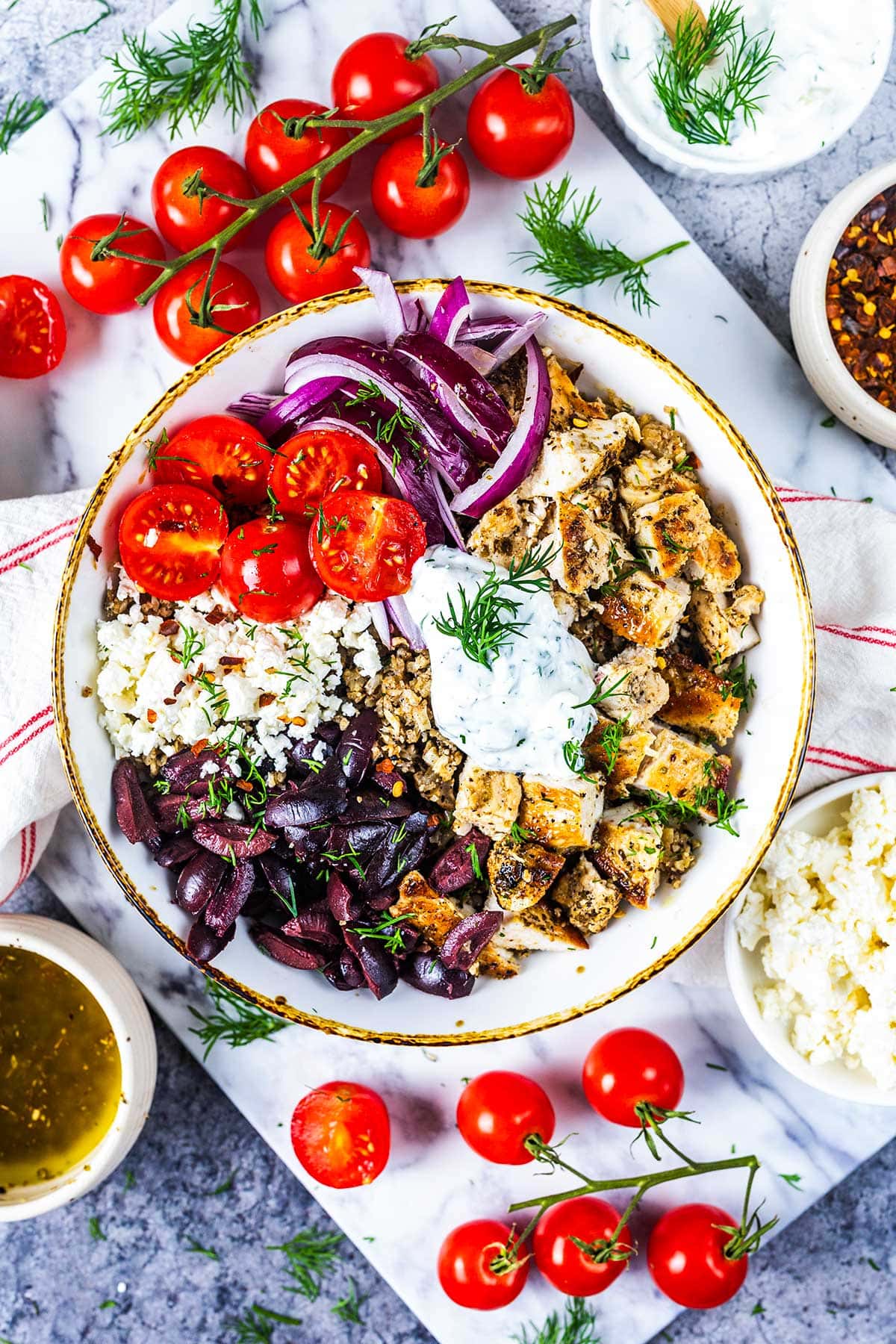 A bowl filled with diced grilled chicken, sliced cherry tomatoes, red onion, olives, cauliflower rice, and tzatziki sauce, surrounded by fresh tomatoes, a bowl of feta, and a small bowl of dressing on a marble surface.