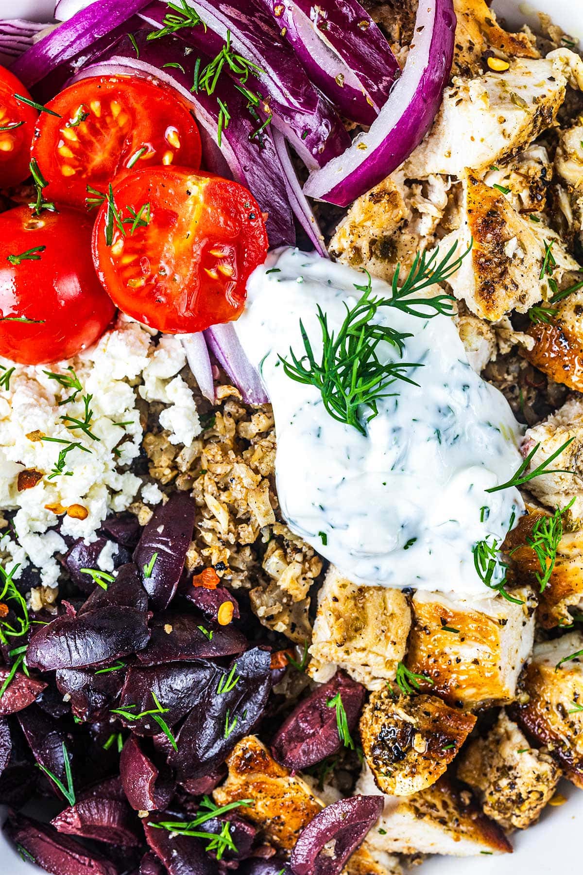 A vibrant Greek bowl with grilled chicken, tzatziki sauce, sliced cherry tomatoes, red onions, Kalamata olives, crumbled feta cheese, brown rice, and fresh dill, arranged in colorful sections.