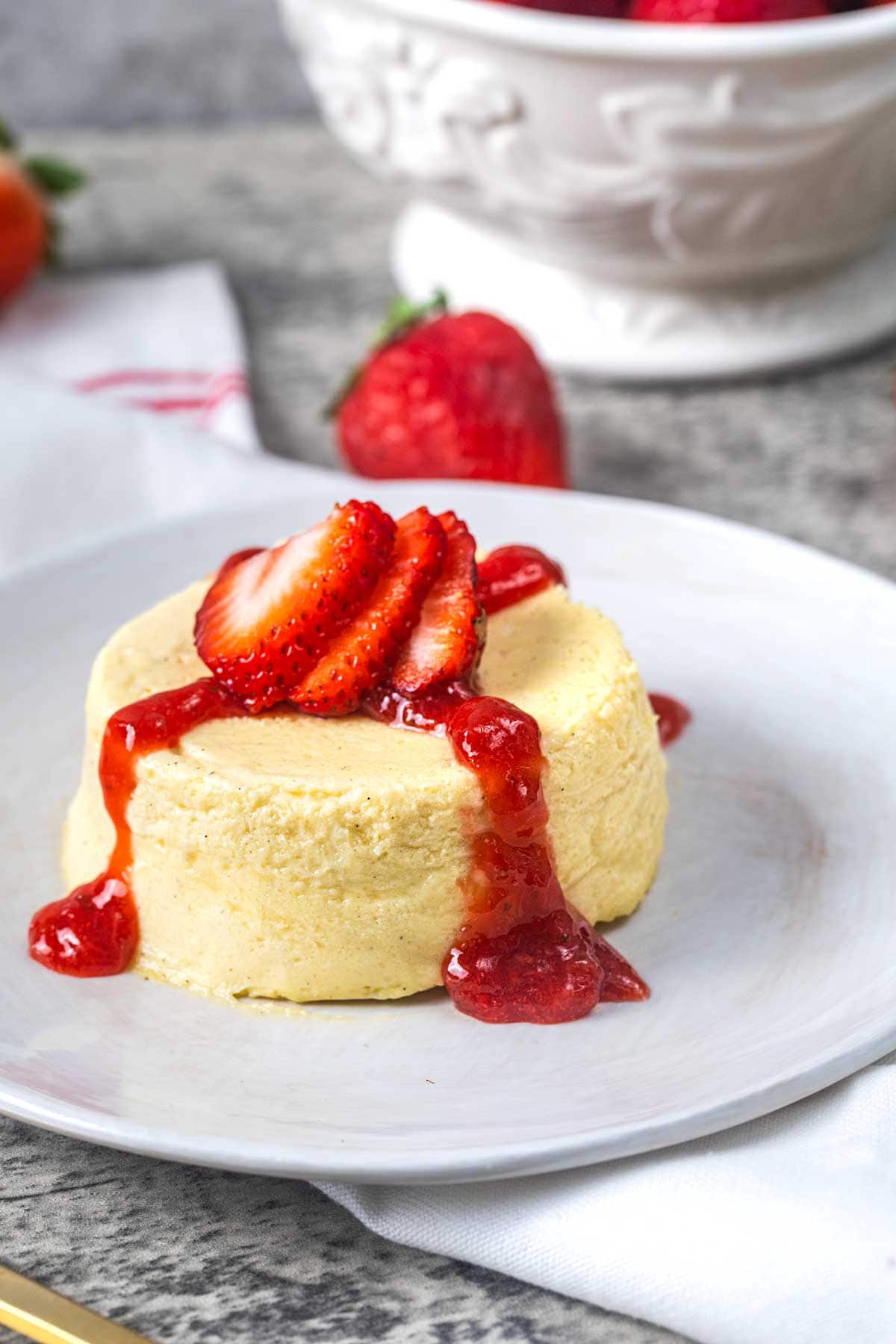 A small cheesecake topped with sliced strawberries and strawberry sauce sits on a white plate. In the background, there is a white bowl filled with whole strawberries.