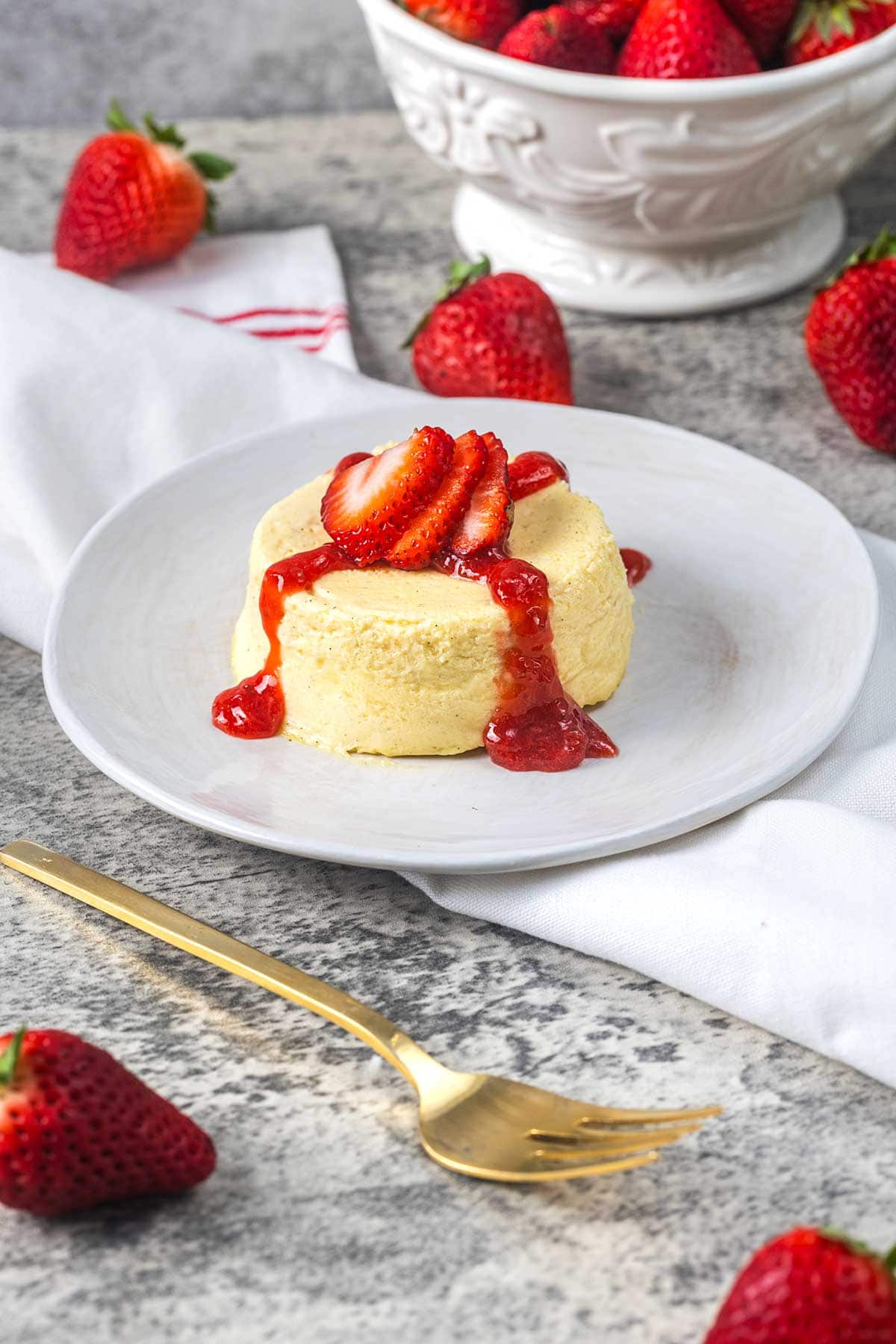 A small cheesecake topped with sliced strawberries and strawberry sauce sits on a white plate, surrounded by whole strawberries and a gold fork. A bowl of strawberries is in the background.