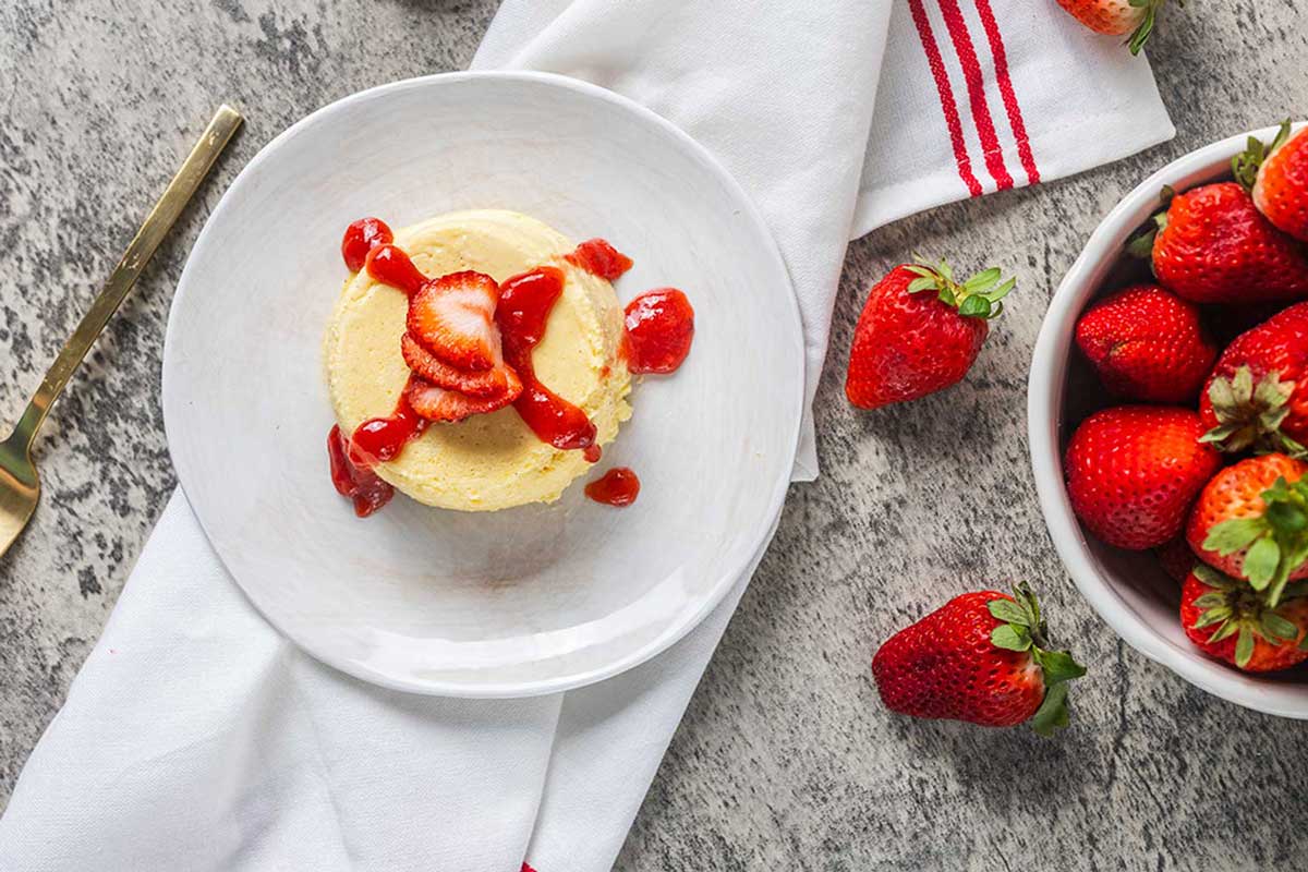 A small keto mug cheesecake topped with sliced strawberries and strawberry sauce on a white plate, next to a bowl of fresh strawberries and a gold fork on a gray surface with a white napkin.