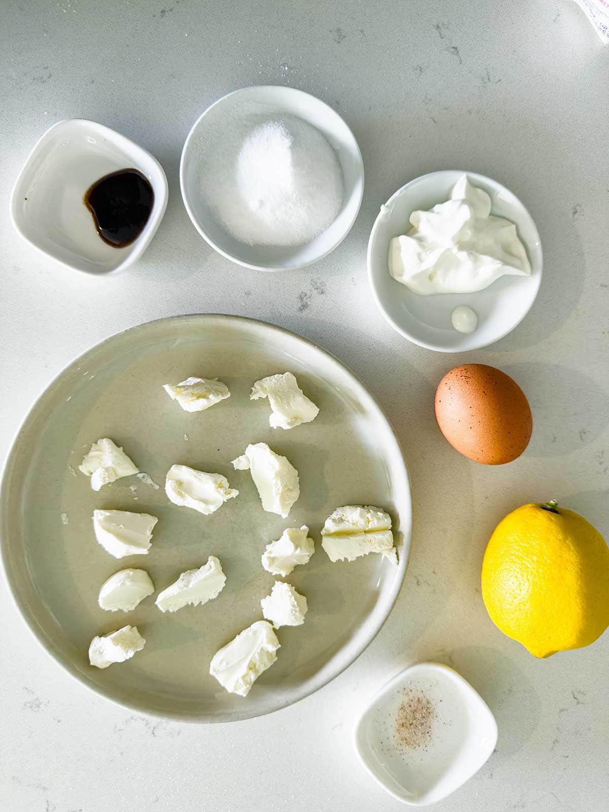 Assorted ingredients on a white countertop: a plate of cream cheese pieces, a lemon, an egg, a small bowl of vanilla, a bowl of white sweetner, a plate of sour cream, and a bowl of salt.