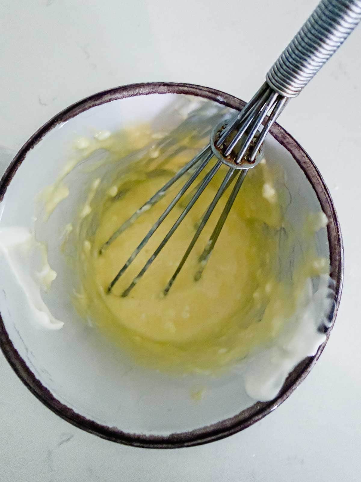 A metal whisk sits in a ceramic bowl containing a creamy, light yellow batter or mixture, with some of the mixture smeared on the inner sides of the bowl.