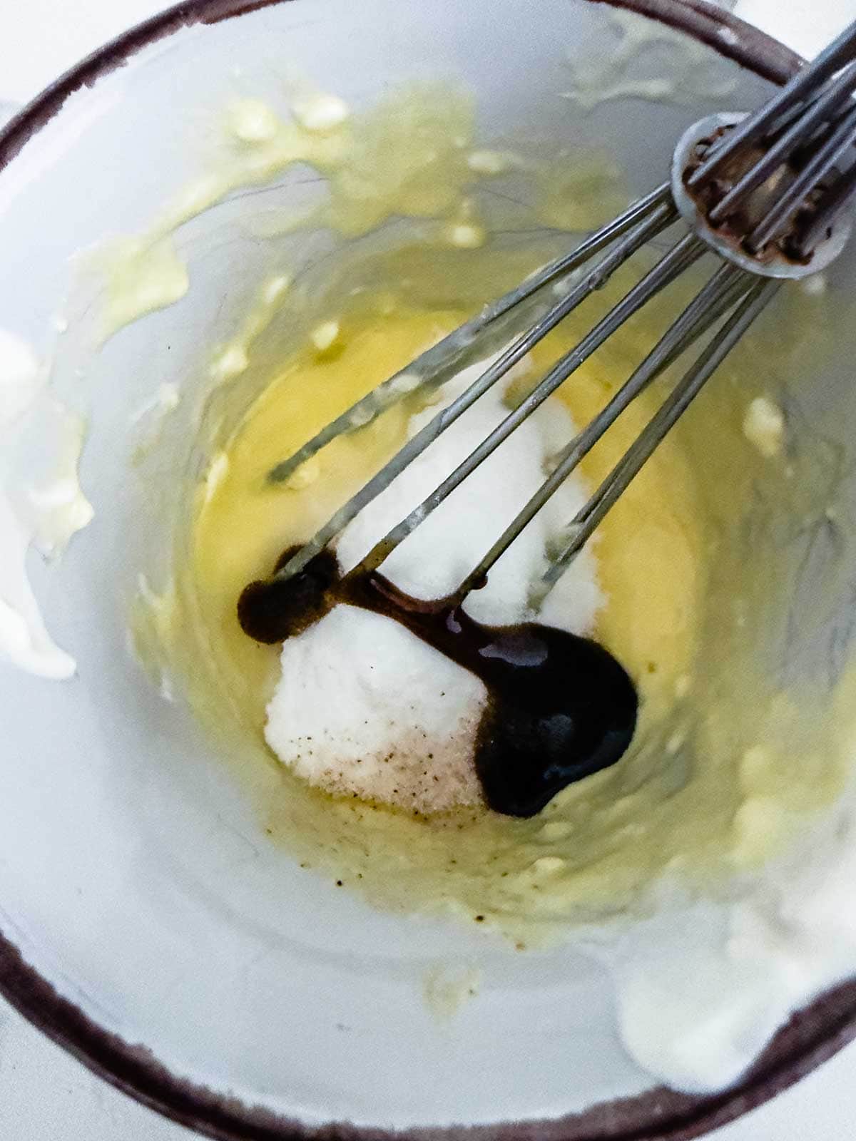 A metal whisk in a bowl with creamy yellow mixture, white sugar, and a dark liquid ingredient, possibly vanilla extract, being combined.