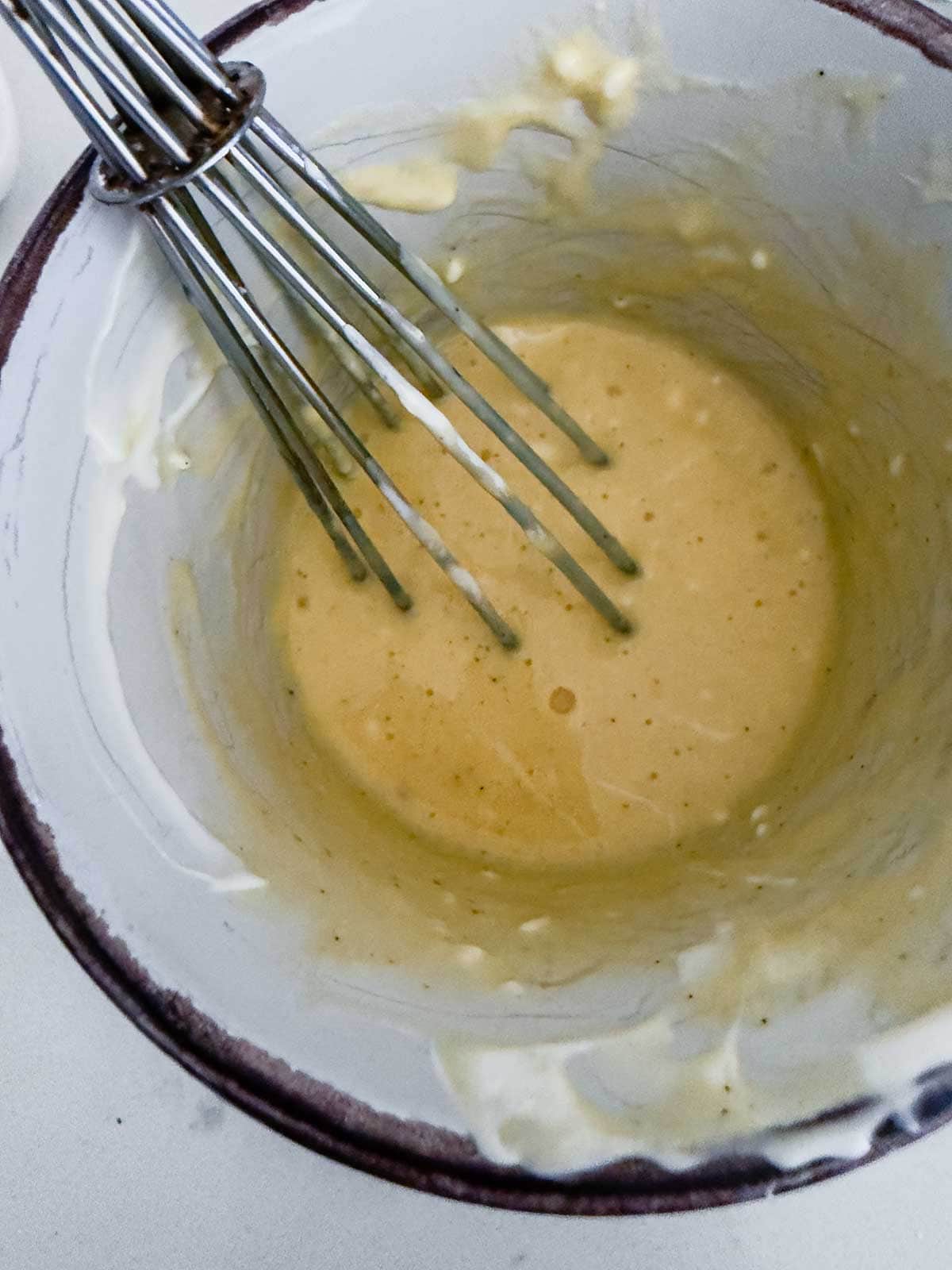 A metal whisk rests in a white bowl with thick, pale yellow batter or sauce partially mixed along the sides. Some residue is visible on the bowl edges.