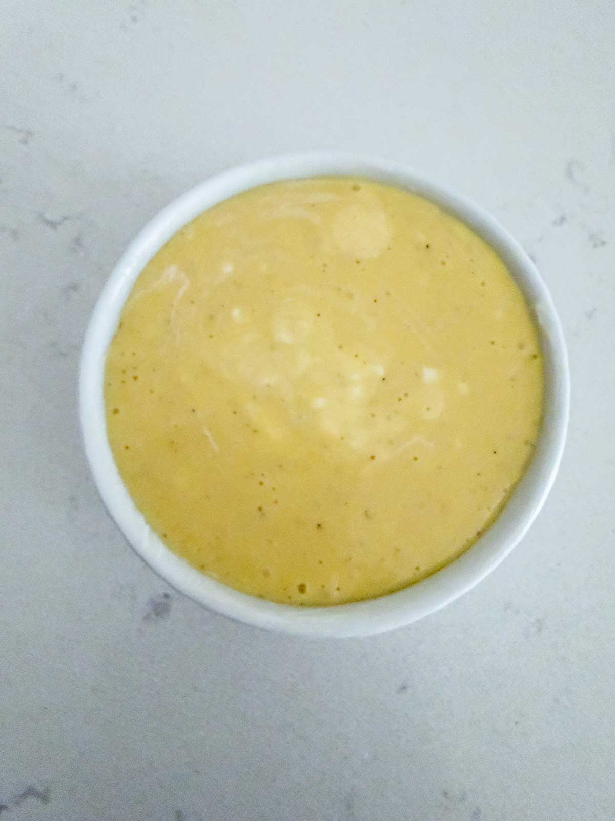 A small white ramekin filled with creamy, light yellow sauce or dip sits on a light-colored countertop.